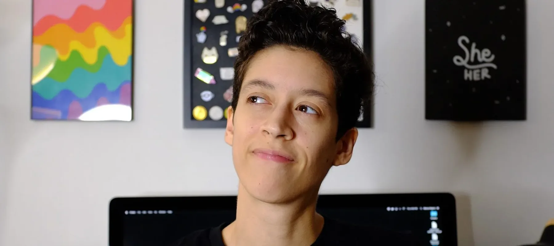 A medium-skinned woman with her short, black hair worn in a pixie cut wears a black shirt with the phrase "Beyond Gender" written on the front surrounded by colorful squiggles. Behind her is a large Mac computer and a few graphic prints hung on the wall.