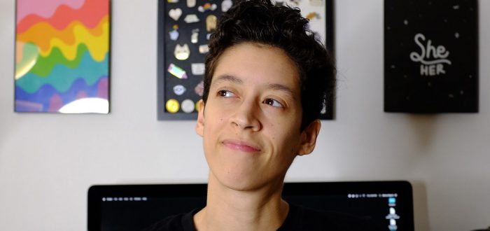 A medium-skinned woman with her short, black hair worn in a pixie cut wears a black shirt with the phrase "Beyond Gender" written on the front surrounded by colorful squiggles. Behind her is a large Mac computer and a few graphic prints hung on the wall.