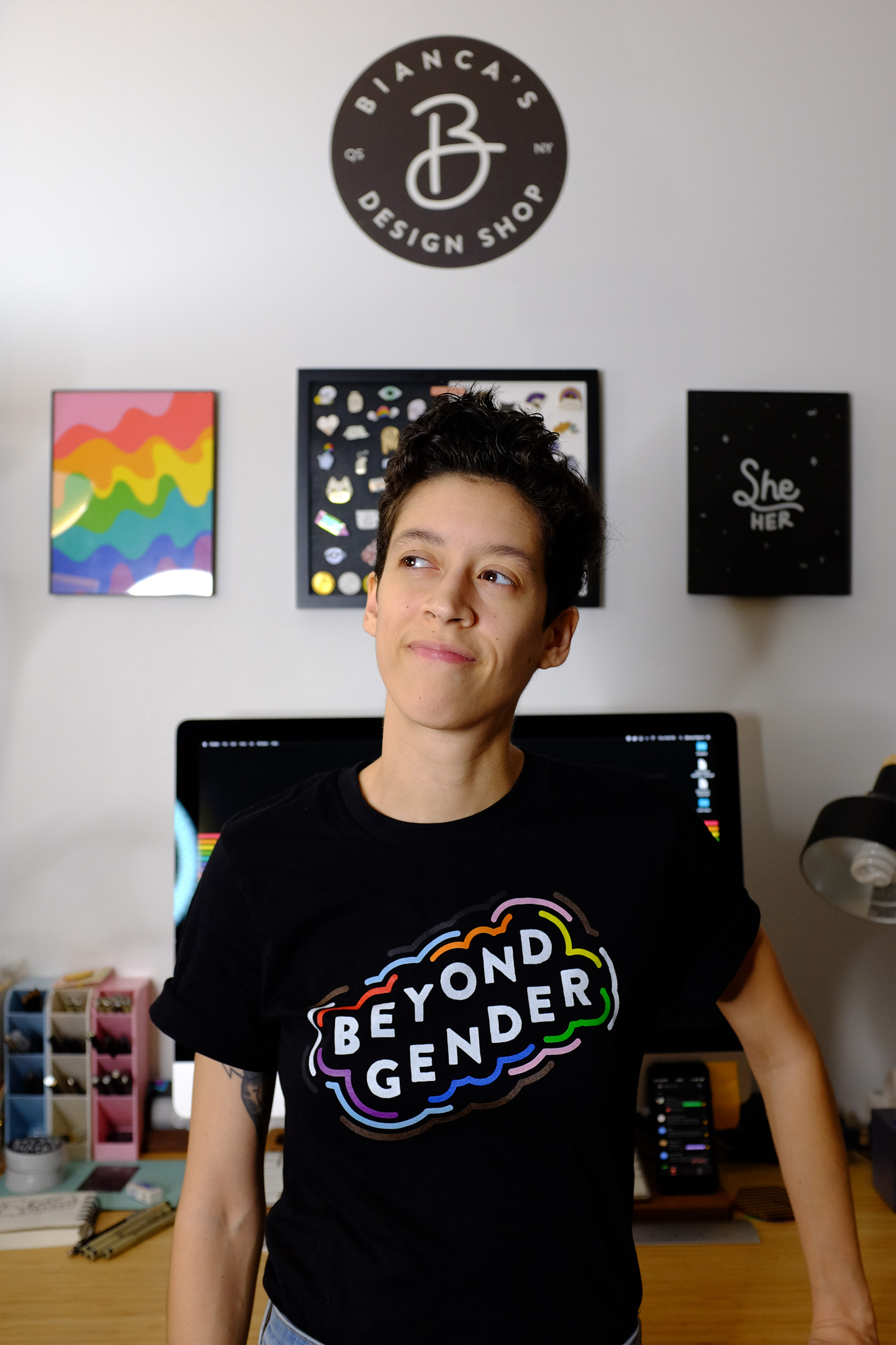 A medium-skinned woman with her short, black hair worn in a pixie cut wears a black shirt with the phrase "Beyond Gender" written on the front surrounded by colorful squiggles. Behind her is a large Mac computer and a few graphic prints hung on the wall.