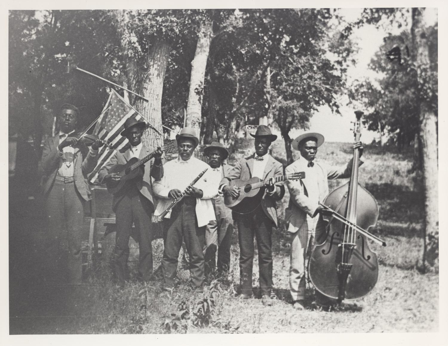 A vintage, black-and-white photo of a group of dark-skinned African American men standing in a wooded area. They are dressed in top hats, suit jackets, and slacks. Most all hold an instrument, including guitars, a cello, violin, and flute. Behind them an American flag is hanging off of a tree.