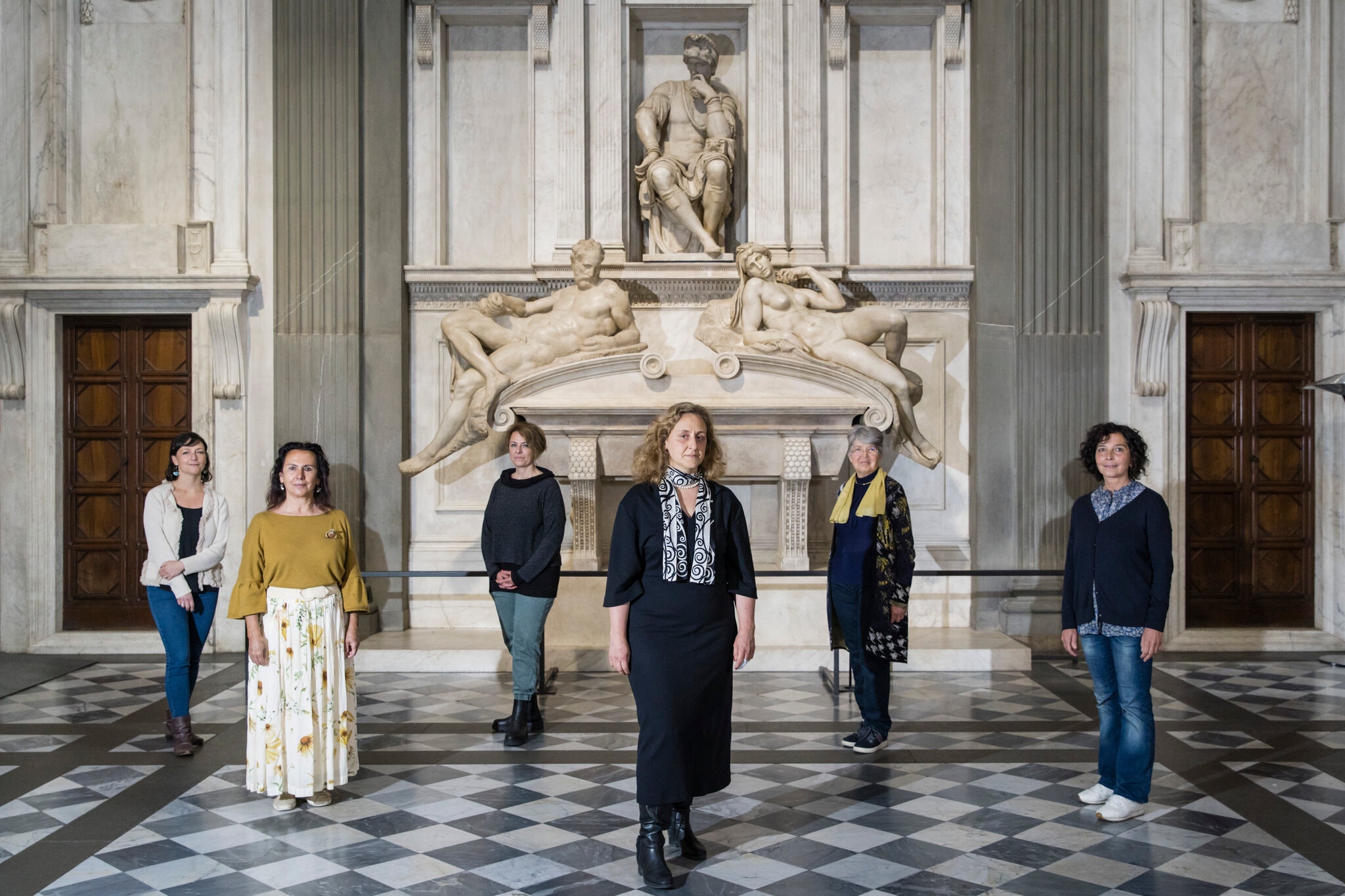 In an ornate, marble hall, with columns and carved statues against the far back wall, six light-skinned woman stand spaced out from one another in a zig/zag pattern. Some wear jeans an sneakers, other are in dresses with boots. They all stare directly at the camera.