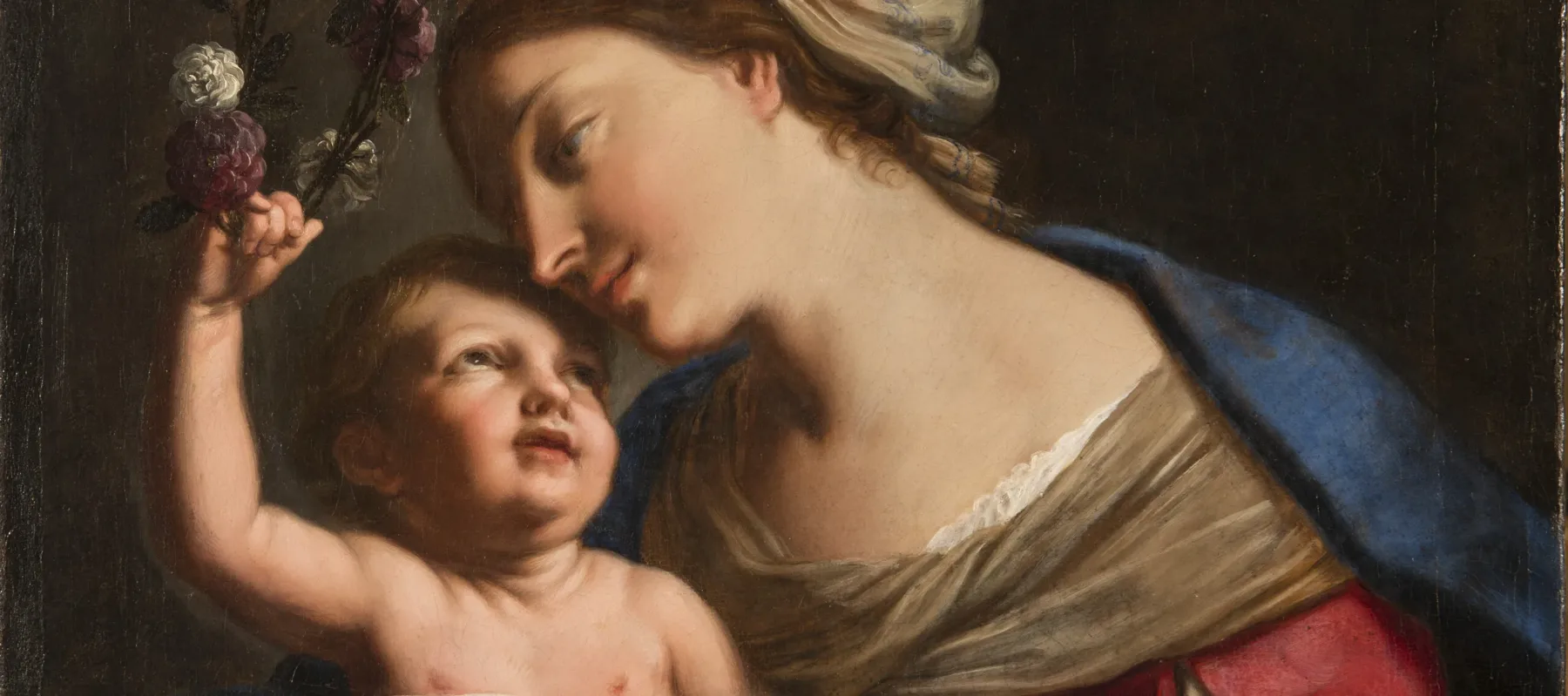 A young, light-skinned, brunette woman gazes down lovingly at the plump baby she holds on her lap. She wears a loose, tan turban, vivid blue cloak, and red dress with white sleeves. The light-skinned child returns her gaze, leaning back to crown the woman with a circlet of roses.