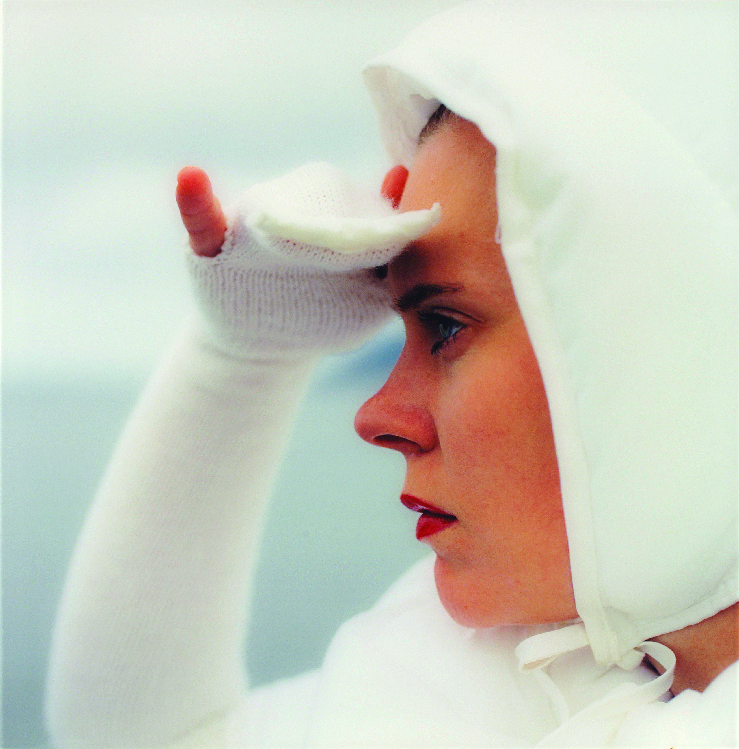 Square close-up of a light-skinned woman in profile looking to the left, seen from the shoulders up. She wears all white, including a bonnet and knit sleeves that become gloves covering her three middle fingers, and she holds her right hand up as if shielding her eyes from the sun.