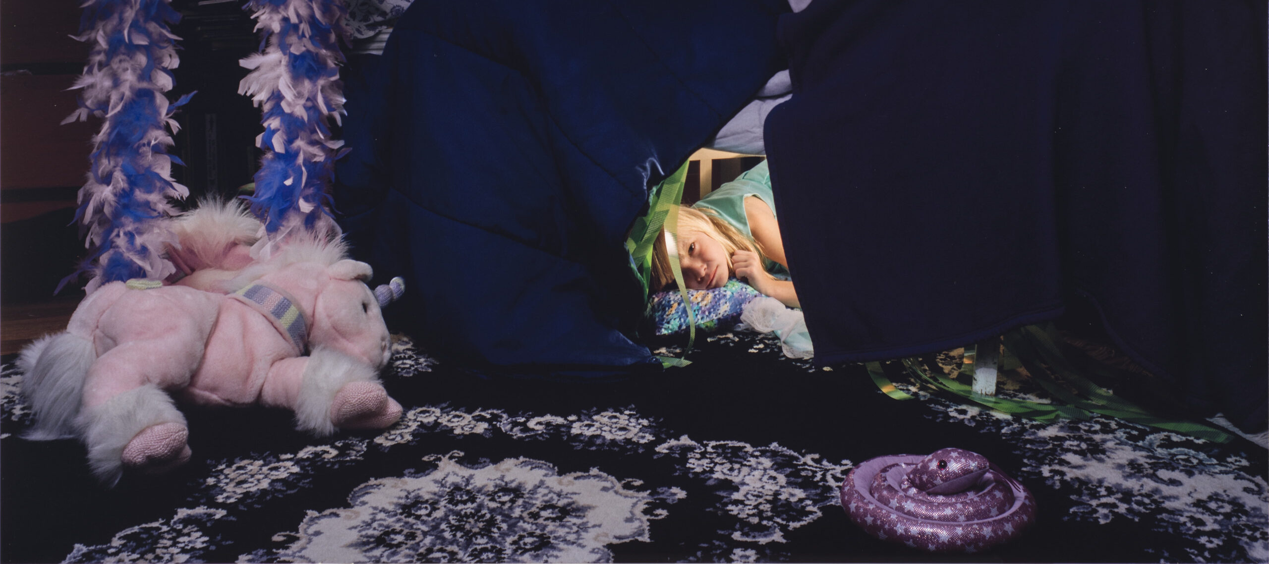 A color photograph of a young light skinned girl with light blonde hair. The girl peeks out from a fort made from a dark blue and purple comforter. A light pink and purple stuffed unicorn sits on a purple and white floral rug in front of the fort. A purple, blue, white, and black paper butterfly-shaped kite hangs on the wall above the fort.