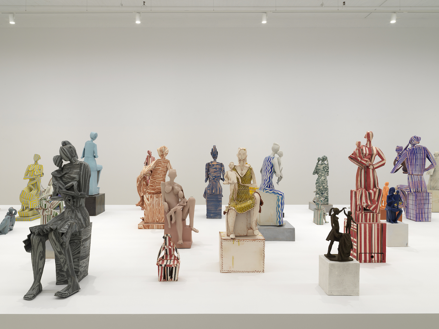 A photograph of approximately twenty abstract Madonna statues on a white table in a sparse gallery. Most are accompanied by a female infant or child. Their bodies are schematic and cubistic. Some figures are nude, some are clothed, some are monochromatic, and some are striped. The statues are all of varying sizes, ranging from under eight inches to three-feet tall.