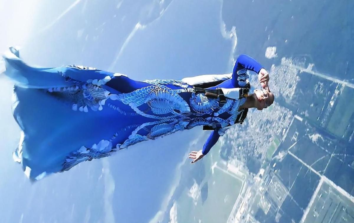 A skydiving woman wears a blue, artistic couture gown as she falls from a plane. She smiles and holds her hand up delicately, looking straight at the camera.