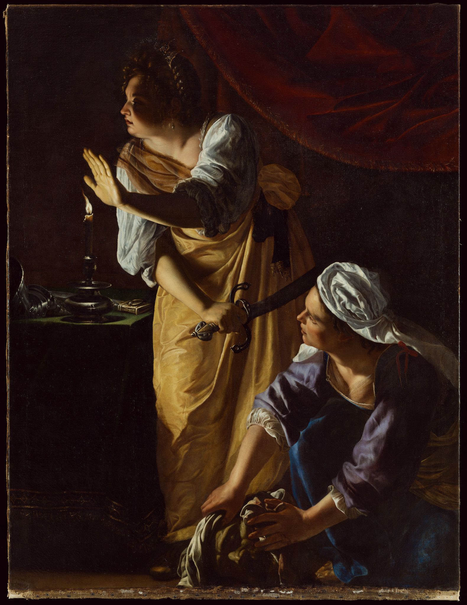 An oil painting from the early 1600s of two light skinned women. One woman stands with her hand outstretched illuminated by a candle on the table next to her. In her other hand she holds a sword with an intricate handle. She is wearing a yellow and white dress and has a jeweled tiara on her head. The other woman is bending down to the floor holding the severed head of a middle aged man in her hands. She is wearing a blue and white dress with white fabric tied around her head.