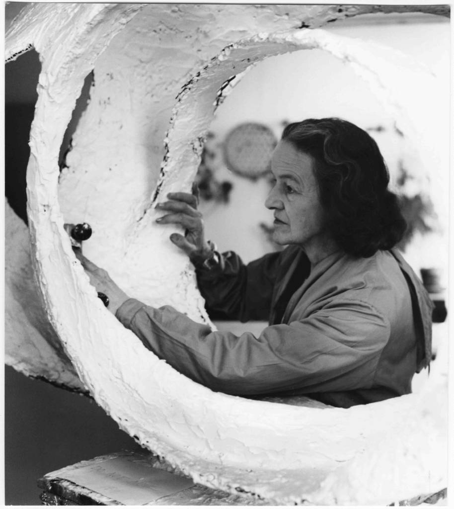 A black-and-white photo of an older light-skinned woman working on a circular plaster artwork. She holds a tool to one side of the work and braces the other side with her free hand. She is focused on the task at hand and stares at the point of work.