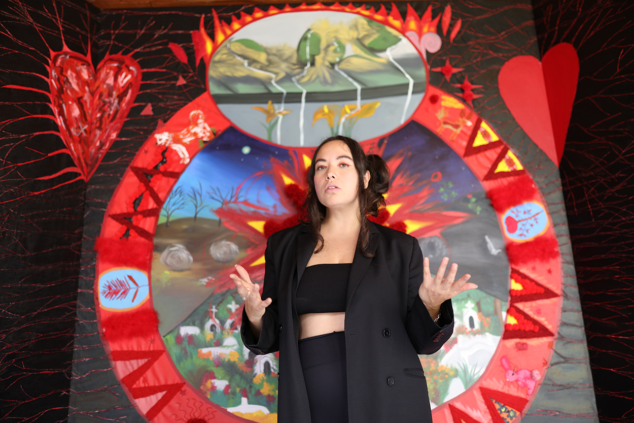 A light-skinned woman with straight dark-brown hair pulled back from her face stands in front of a large painting. She wears a black tube top, leggings, and oversized black blazer. Her hands are in motion, as if she is mid-conversation. The painting features two red hearts, one smooth the other painted with erratic lines, on either side of a mandala featuring different sections of imagery, including nature scenes.