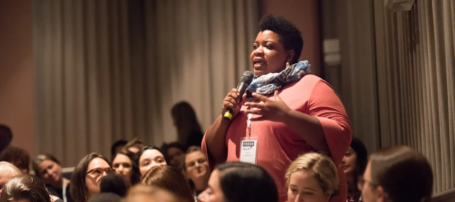 A person with dark skin and short black hair stands amongst a seated audience and speaks into a microphone.