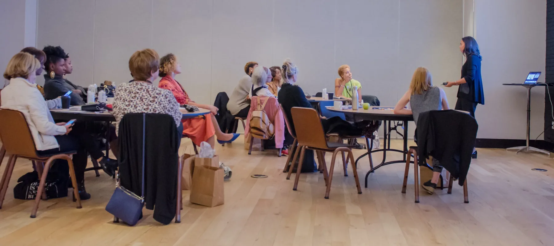 A large group of women are seated randomly in a large room listening to a speaker.