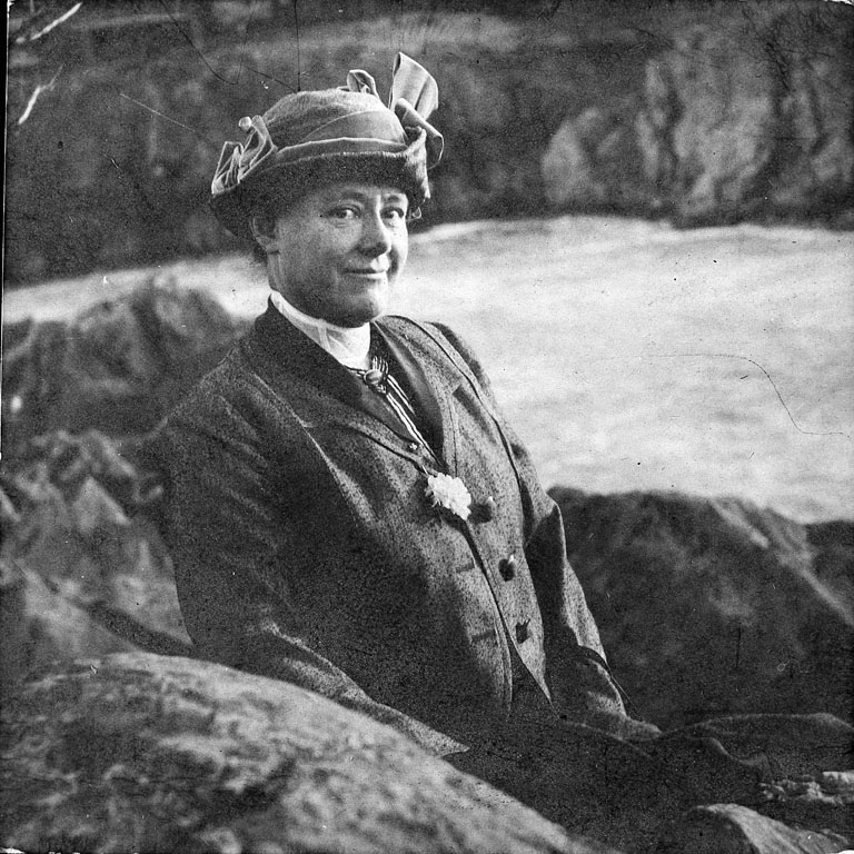 A black-and-white photo of a light-skinned woman from the early 1900s posing between large rocks with a river behind her. She wears formal dress and a hat and smiles slightly at the camera.