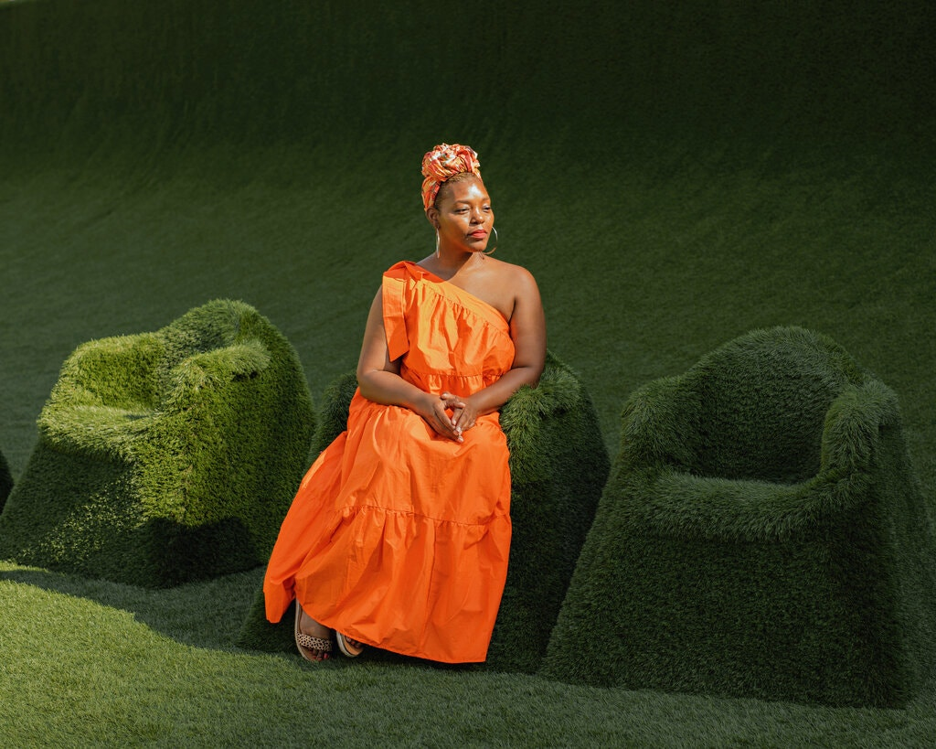 A dark-skinned woman wearing an orange one-shoulder dress and headwrap sits in a regal, contemplative pose in a chair fully covered in dark green grass. There are two other unoccupied chairs on either side of her, and the background of the image is also grass. The sun shines on her