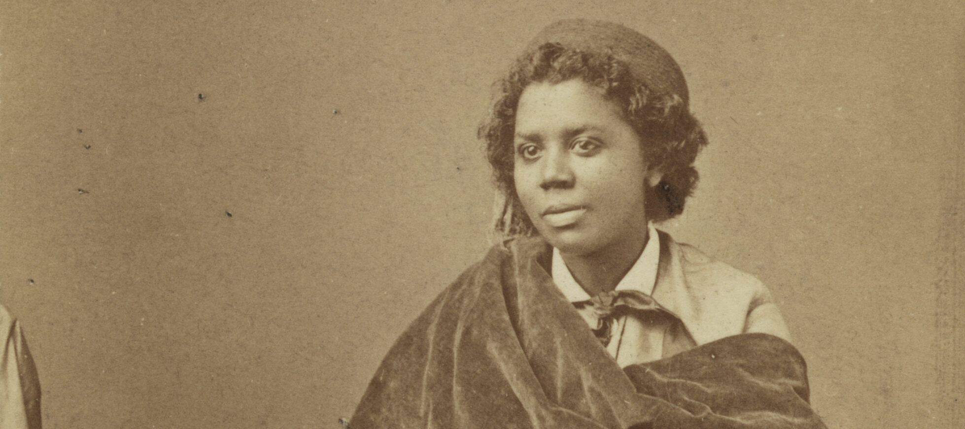 A black and white photograph from the late 1800s of a dark skinned woman seated on an upholstered chair with fringe around the arm and seat. She is wearing a white collared shirt with a scarf tied around her neck, a light colored jacket and a matching ankle length skirt. There is heavy, dark colored velvet fabric draped from her right shoulder to her left arm. She is wearing a round, flat-topped hat over her short wavy hair.