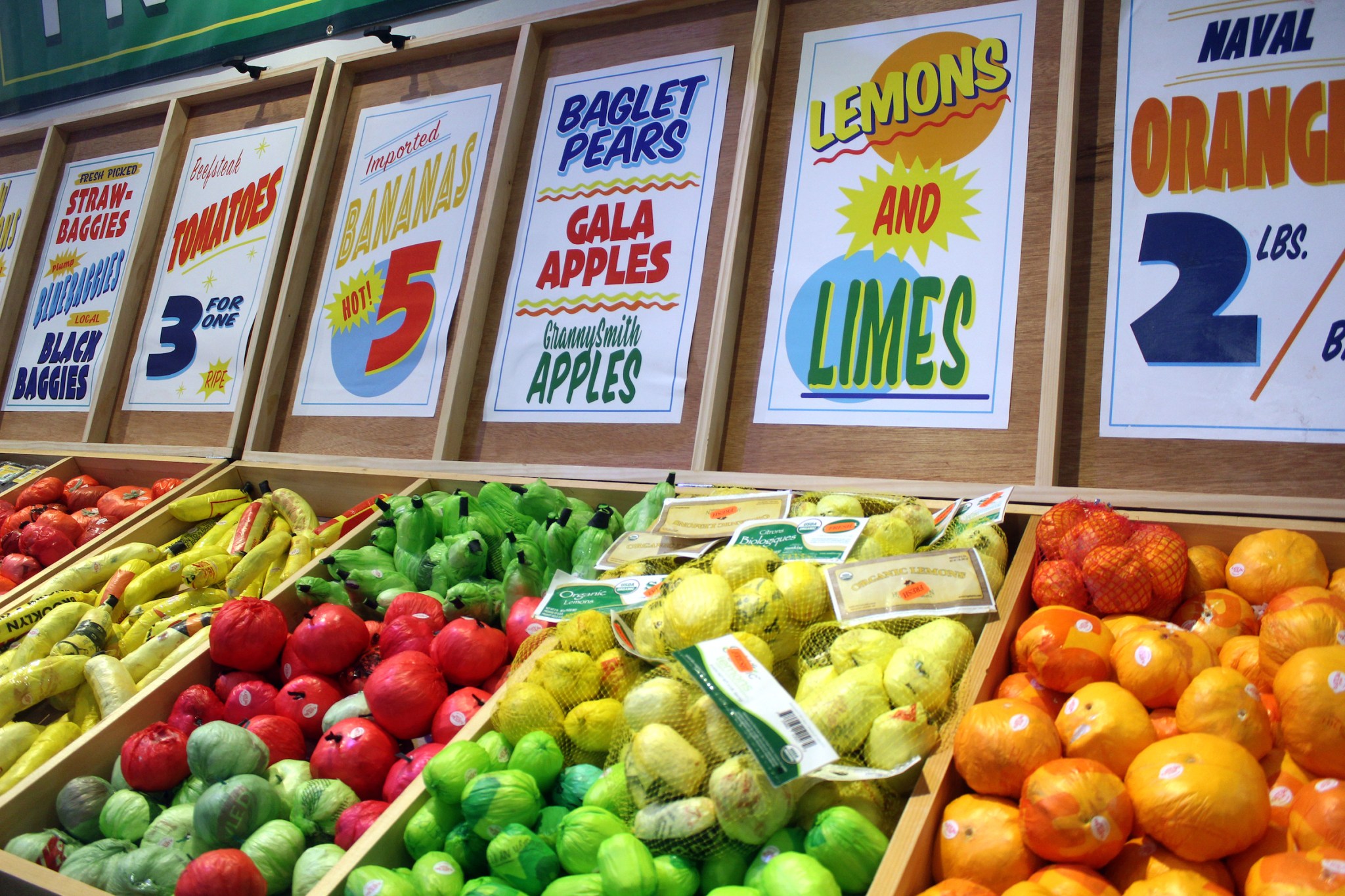 A color photograph shows four wooden grocery store produce bins filled with bananas, apples, lemons and limes, oranges, and more. Above each bin a colorful sign notes the fruit's name in colorful, fun lettering.