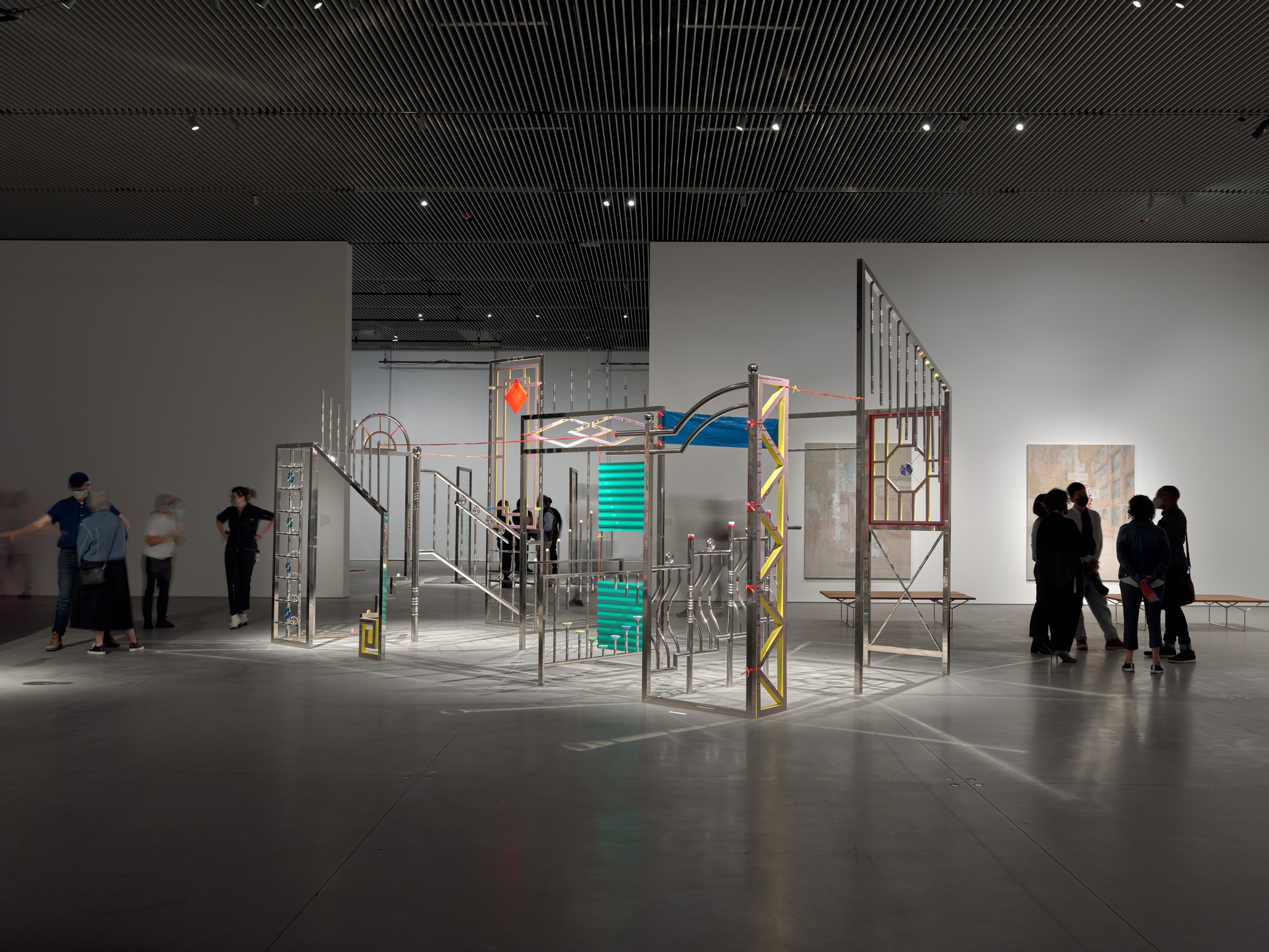 In a sparse gallery, large stainless steel rods are tack welded to resemble staircases, doorways, and windows, complete with architectural decorations. The majority of the sculpture is silver, though some of the decorative motifs are yellow, pink, orange, purple, or blue. About a dozen gallery-goers stand next to and behind the installation.