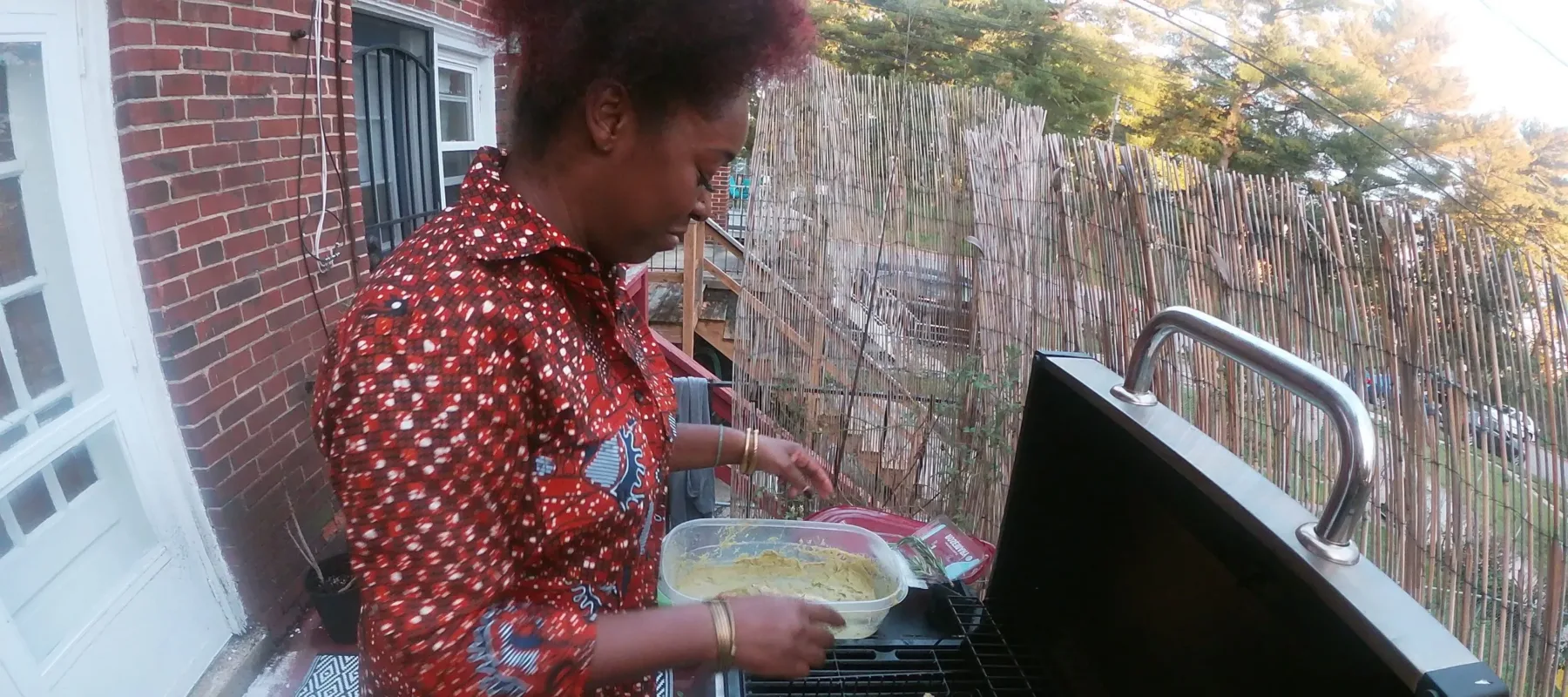 A dark-skinned woman stands on an outdoor balcony, grilling four whole fish on a gas grill. She holds a Tupperware of marinade. The photo is taken with a GoPro, creating a fish eye angle of the scene.