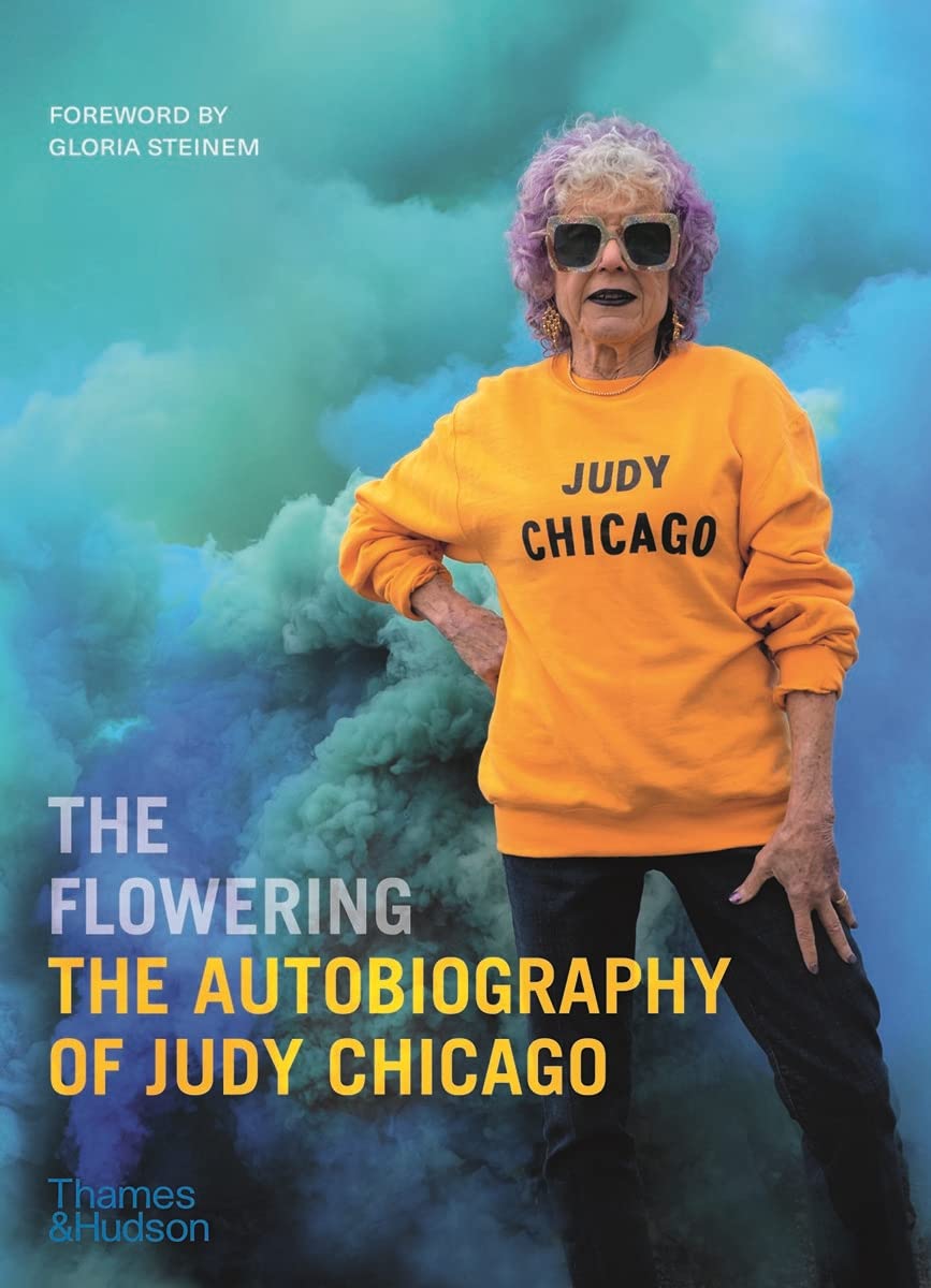 A book cover features bright blue smoke plumes. A light-skinned older woman with purple, curly hair stands with one hand on her hip. She wears a golden-yellow sweatshirt that says "JUDY CHICAGO" in black letters. Atop the book cover, "The Flowering: The Autobiography of Judy Chicago" is written.