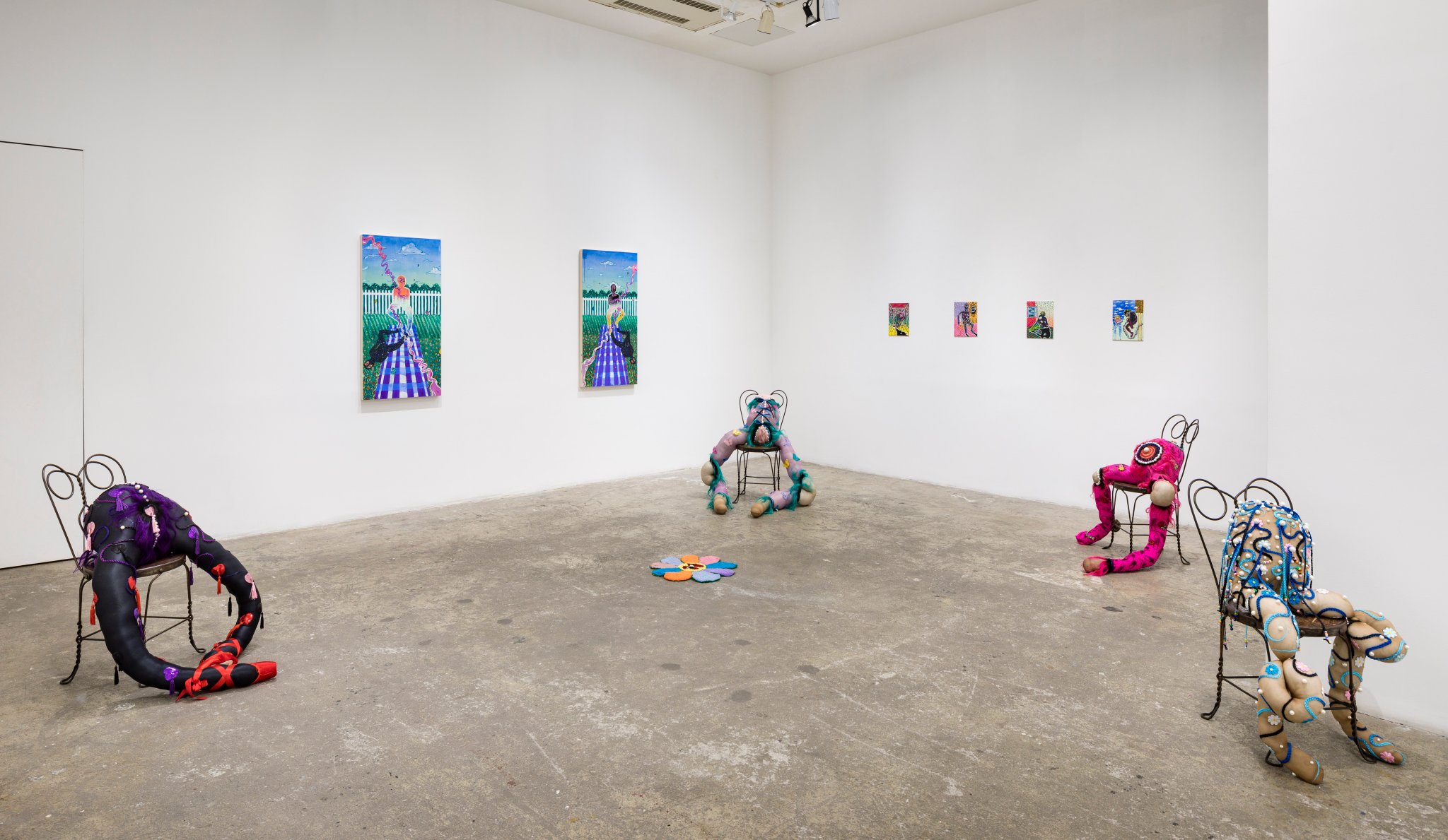 An installation photograph of abstract sculptures and paintings in a sparse gallery. Four small and two large colorful paintings hang on the walls. Four anthropomorphic soft sculptures are each seated on garden chairs, and their lanky and lumpy limbs are adorned with pearls, sparkles, colorful braids, and ribbons. A sculpture of a flower rests in the middle of the floor.