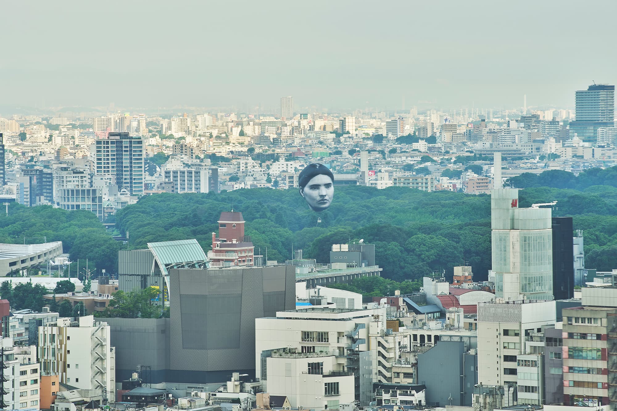 A photo of the Tokyo skyline on a hazy day. In the mid-ground, a sea of green treetops divides the cityscape. Above the trees floats a large balloon face with a black-and-white portrait of an anonymous person. The subject has dark hair parted in the middle, dark eyebrows, and a serious expression.