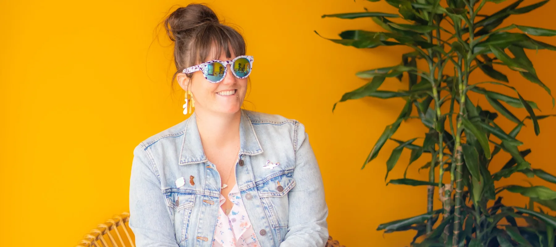 A light-skinned woman sits in a circular rattan chair in front of a bright yellow wall. She wears sunglasses, a jean jacket, a light patterned dress that hits above the knee, and blue sneakers. She smiles off into the distance. Next to her is a tall tropical plant in a terra cotta pot.