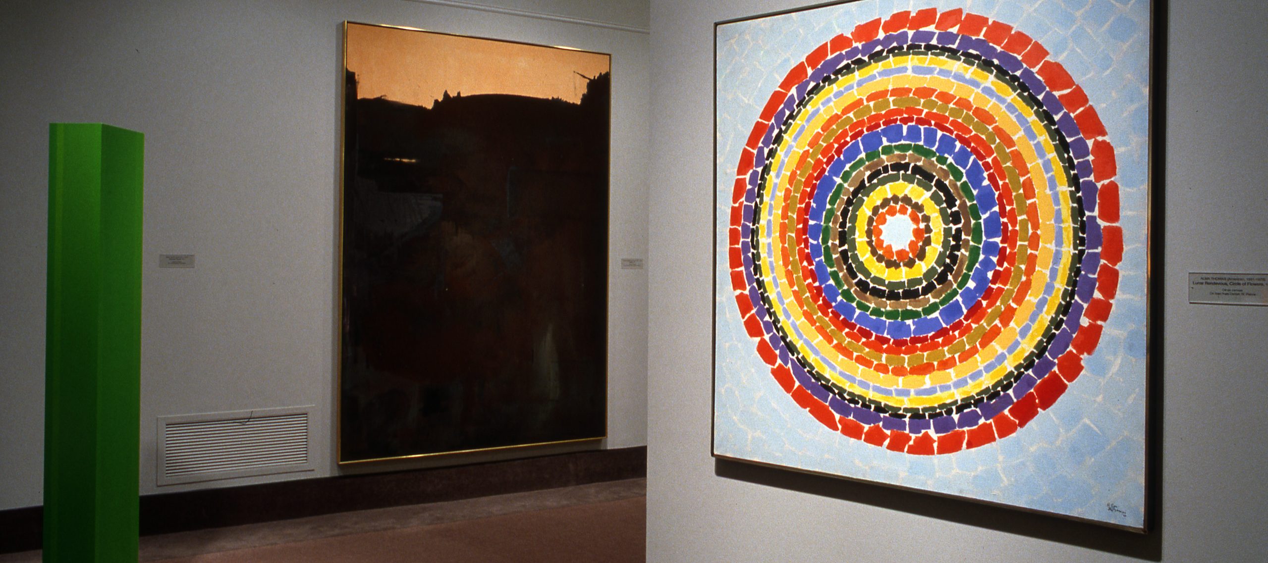 Gallery view of three artworks: a tall green column on the left, a multicolored painting of many concentric circles on the right, and a large black-and-white painting in the background between the two.
