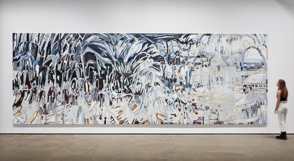 In a white-walled gallery, a light-skinned woman with light brown hair dressed in a black tank top, white pants, and black boots stands to the right of a large, horizontal abstract painting. The canvas features gray, silver, light brown, and tan thick lines painted in waves up and down the canvas. As the canvas moves from left to right, the palette grows progressively lighter in color.