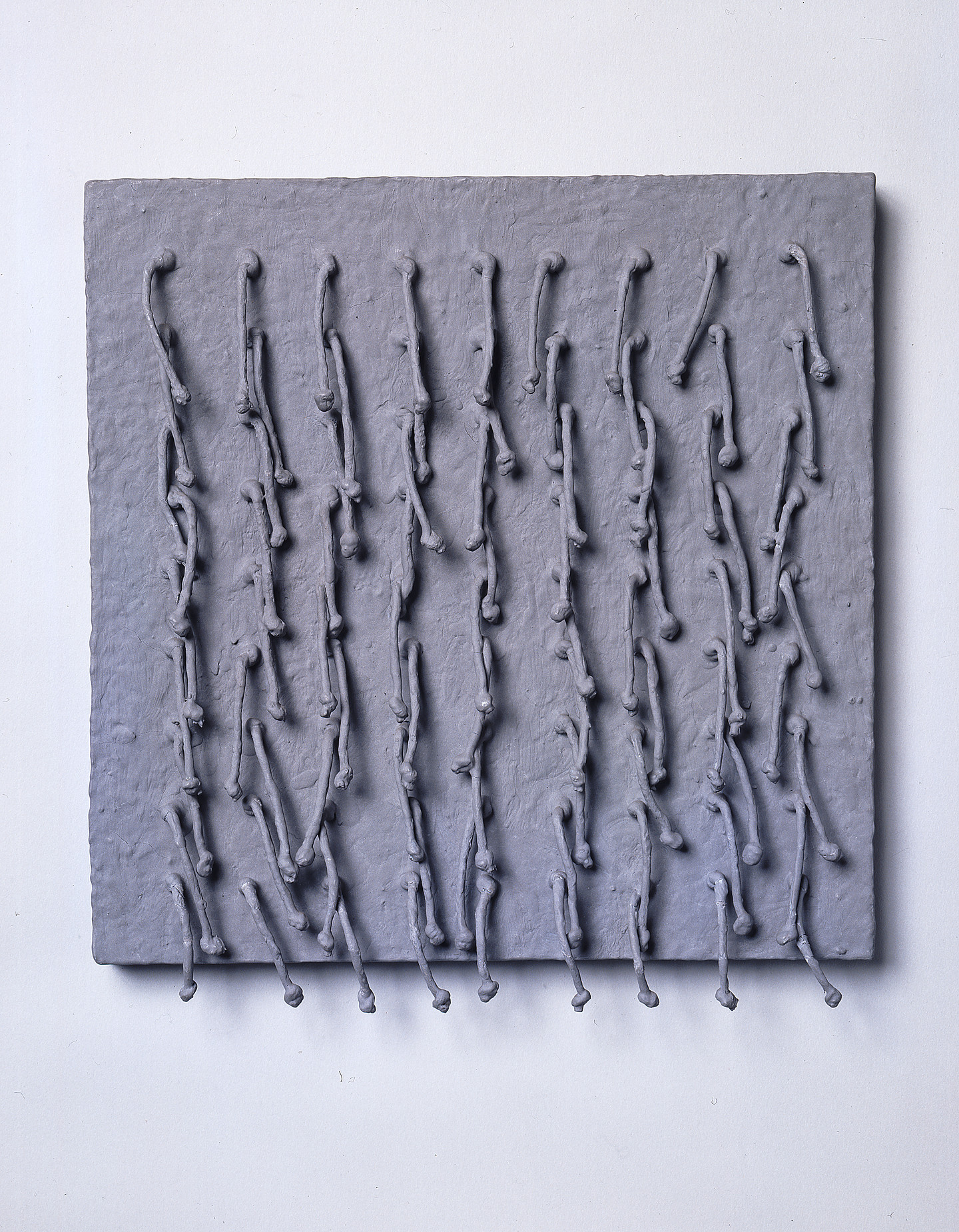 Short, knotted cords, arranged in a symmetrical 9-by-9 grid emerge from a square panel and hang down. Thick, matte-gray paint coats the entire piece, adding rough texture to the panel and stiffening the cords into positions that cast irregular shadows across the work’s surface.