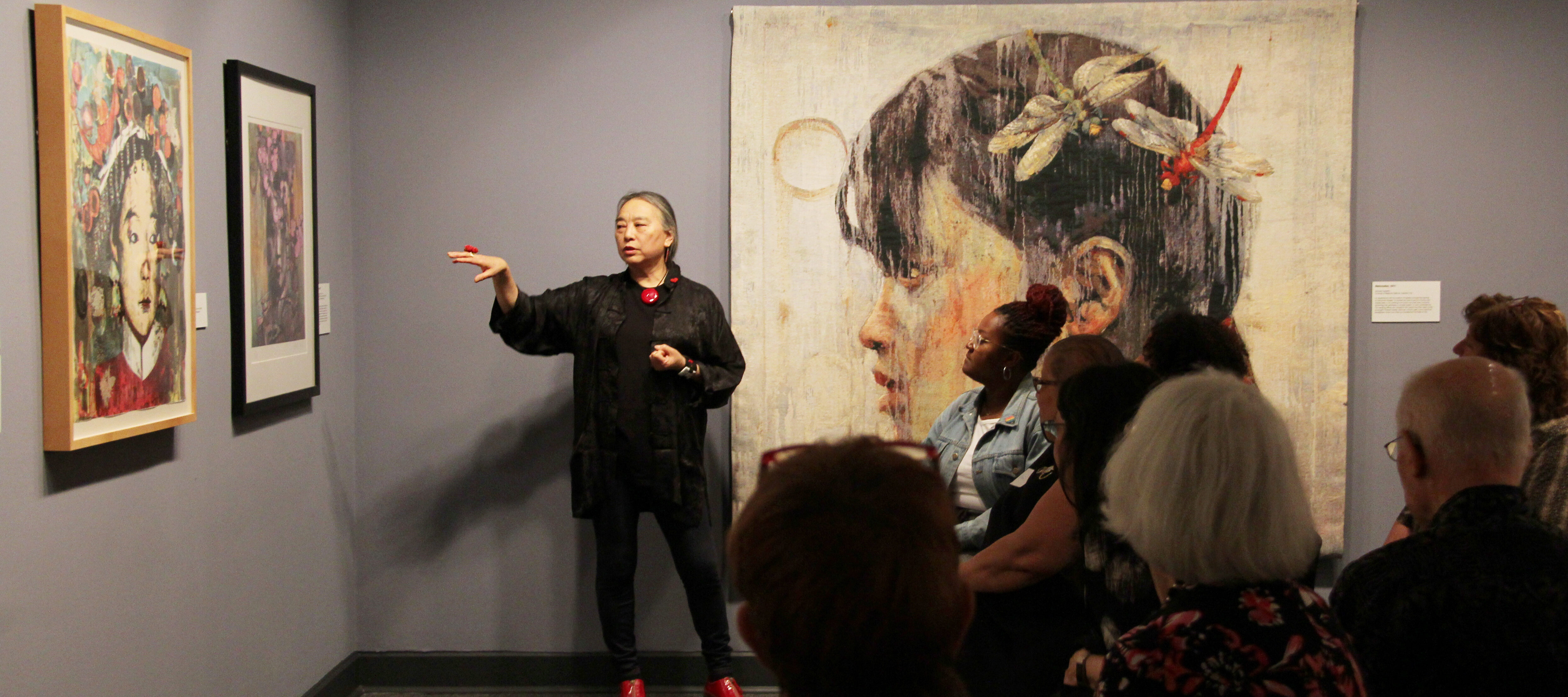 Hung Liu speaks to an audience about her paintings of Chinese women, which hang on the walls around her. Liu is a light-skinned adult woman with gray hair in a low ponytail, dressed in black with red jewelry and shoes. She stands, gesturing with one hand at a painting to her right.