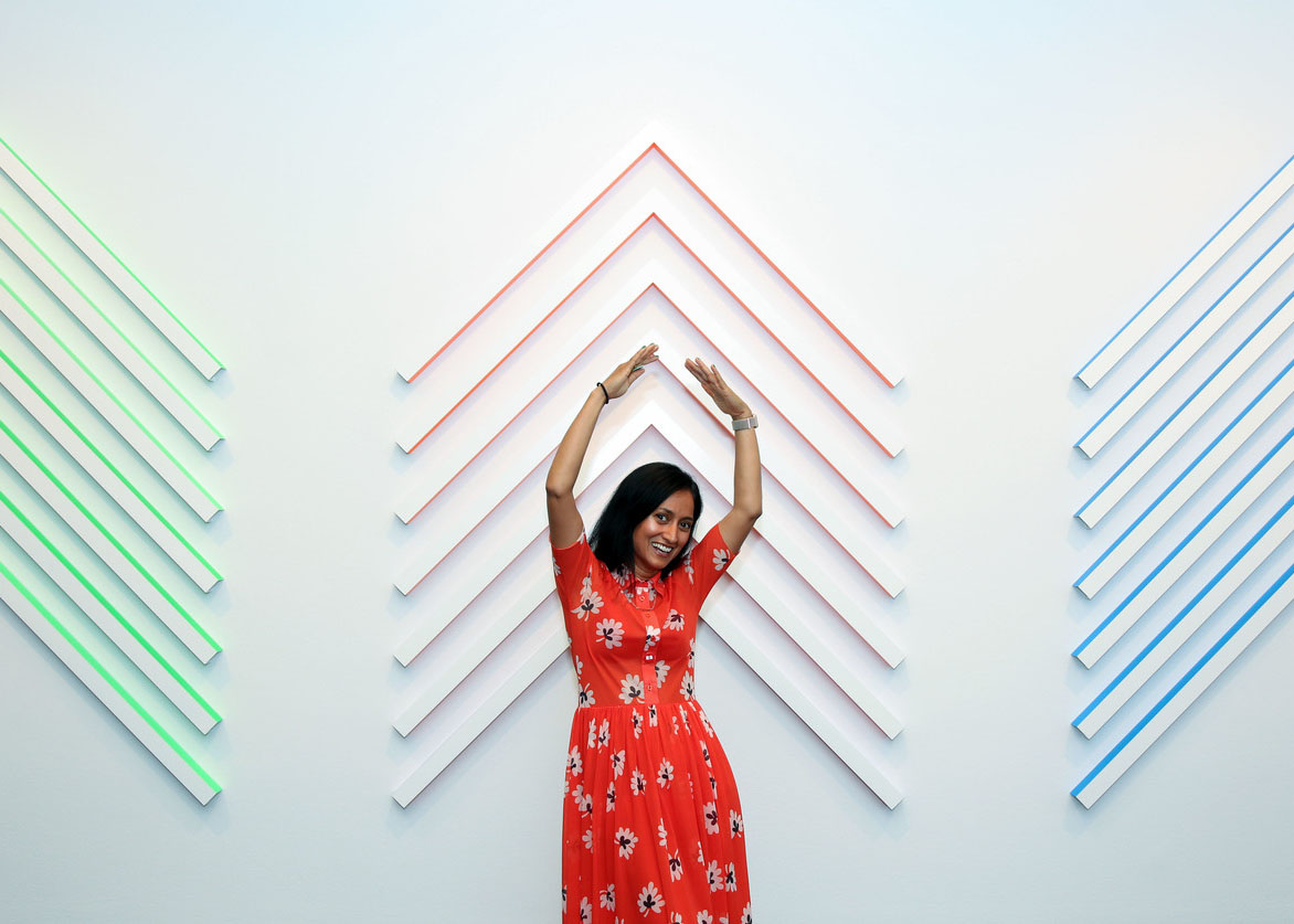 A photograph of a medium-skinned woman with a smile on her face, wearing a red dress with white and black flowers. She stands in front of a geometric wall sculpture with her hands stretched above her head.