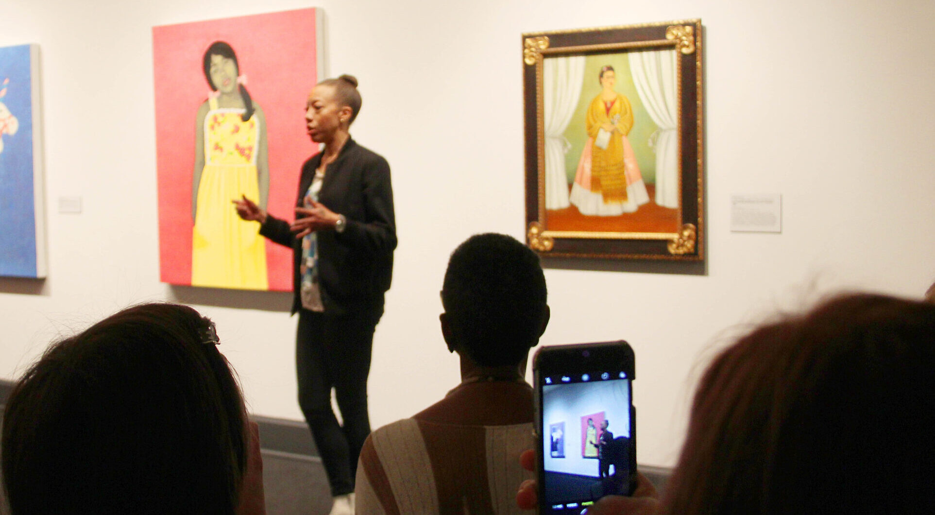 Smartphone filming of a darker skinned woman artist talking in front of her pink and yellow artwork in the museum gallery.