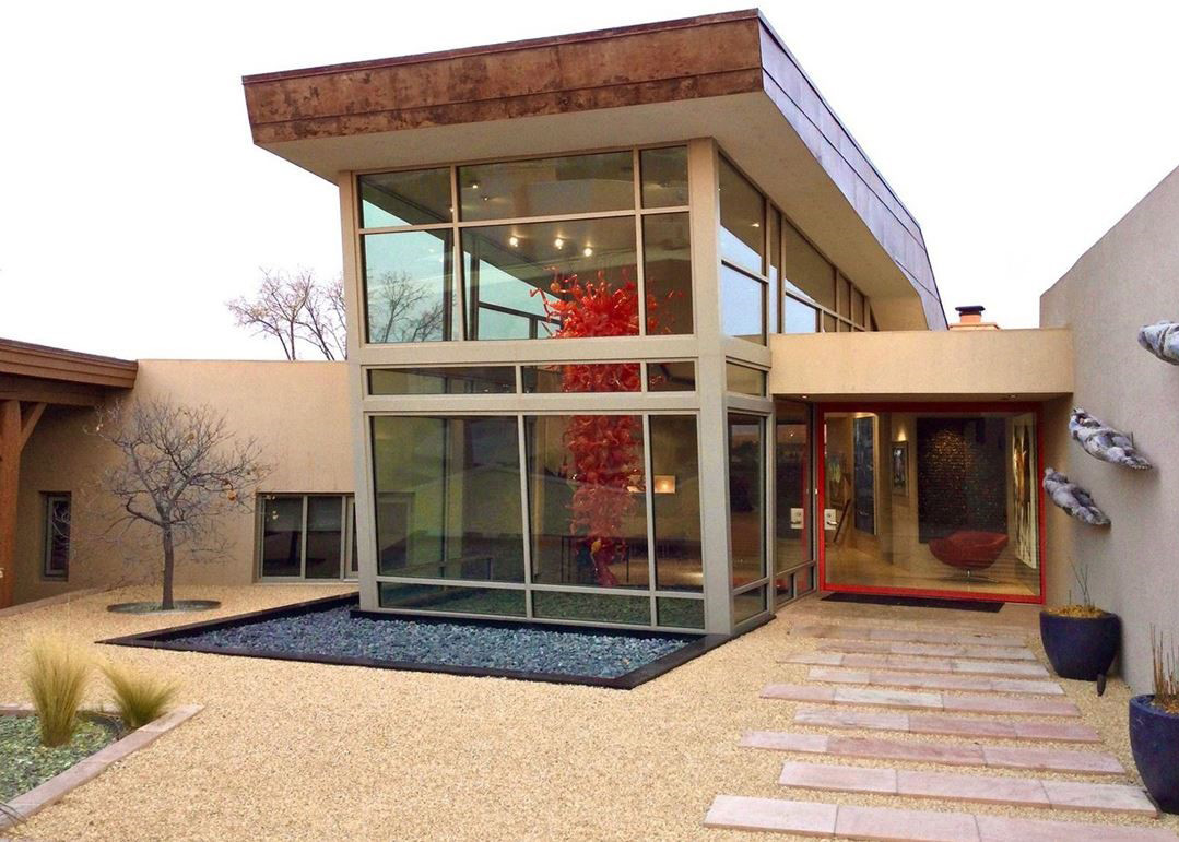 A photograph of a modern style house with a two-story window at the entrance. There are three metal sculptures along the outside wall and a large sculpture hanging from the ceiling inside made of many pieces of curved blown glass.