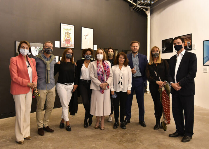 A group of 10 light-skinned men and women stand in front of a wall of artwork. The figures are dressed in smart casual attire and are wearing face masks.