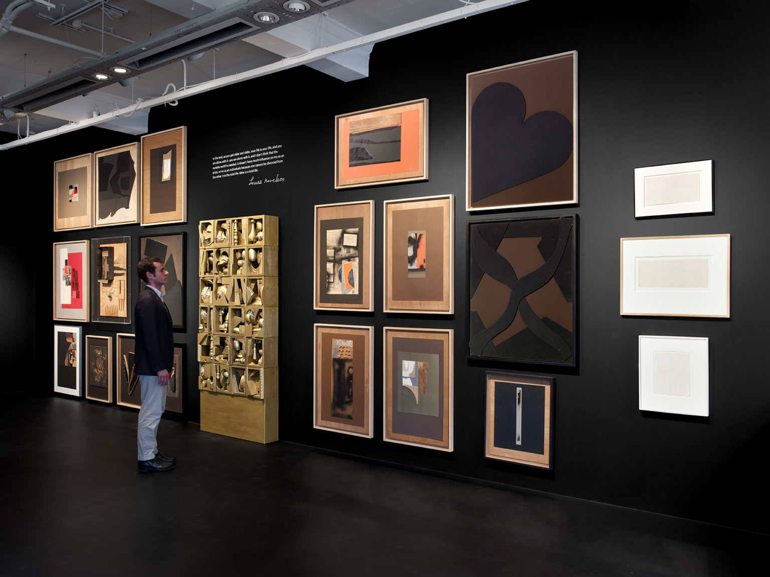 In a gallery with a black floor and walls, a light-skinned, brown-haired man wearing a blazer stands before a wall showcasing 22 of Louise Nevelson’s artworks. In the center of the wall stands s sculpture that resembles a bookshelf of knickknacks. A quote by Nevelson is written on the wall space above the bookshelf. Hanging on the walls flanking the bookshelf are several abstract collages and works on paper.