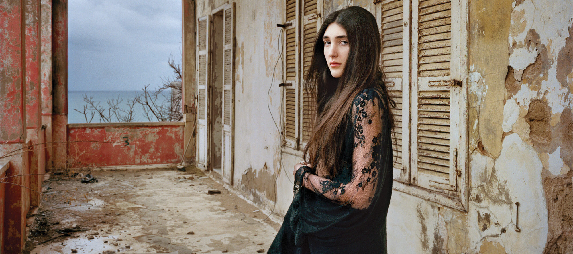 A light-skinned young woman with long, dark brown hair in a black, long lace sleeved dress stands confidently in a crumbling loggia. She gazes at the viewer with a serious, captivating look.