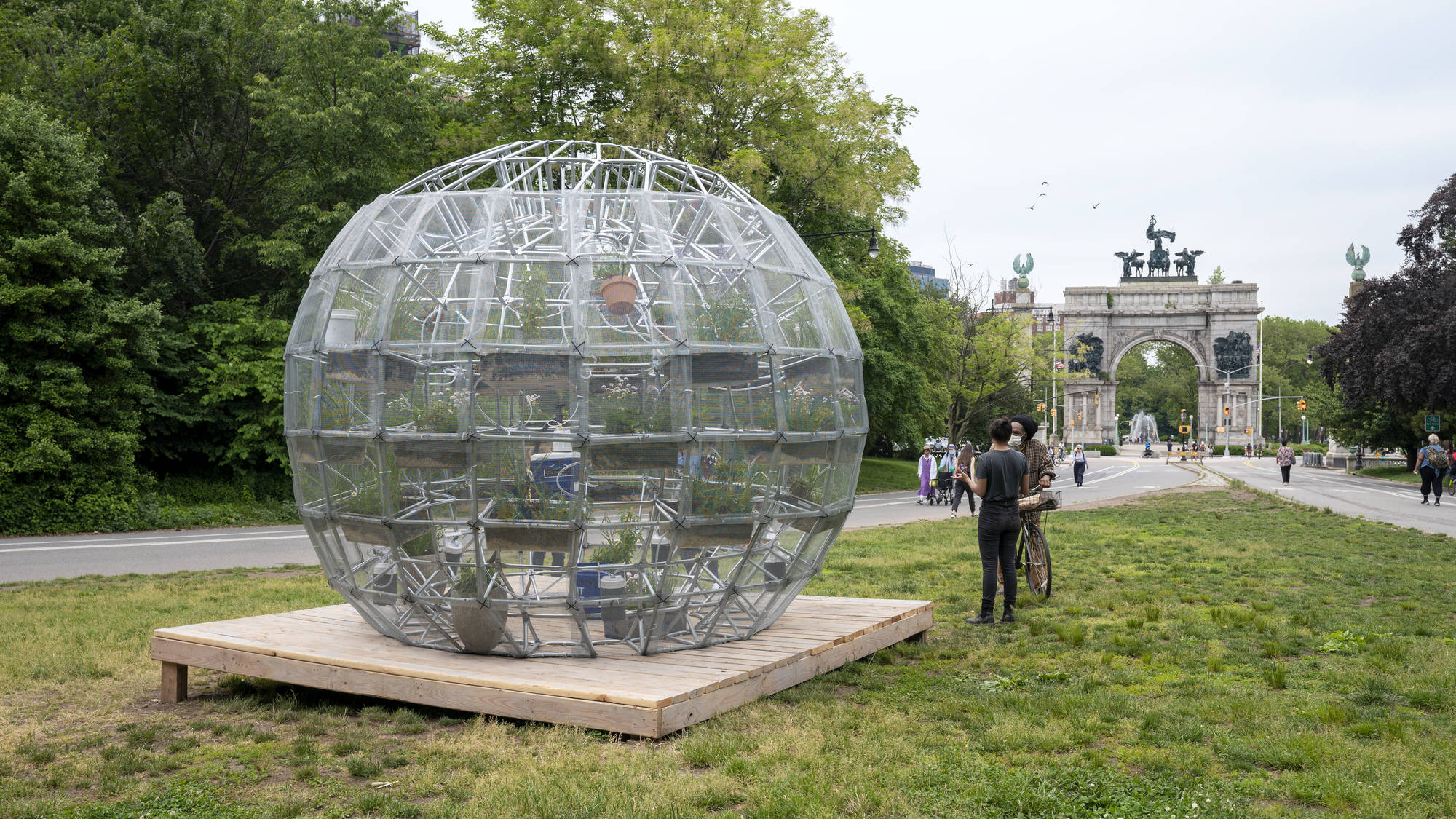 A photograph of a park on a cloudy day. On a strip of green space is a 10-feet-tall geodesic dome sitting atop a plain wooden platform. Inside the dome is an assortment of potted and hanging plants. Two men, one light-medium-skinned and one dark-skinned, stand to the right of the dome and gesture as if discussing the sculpture.