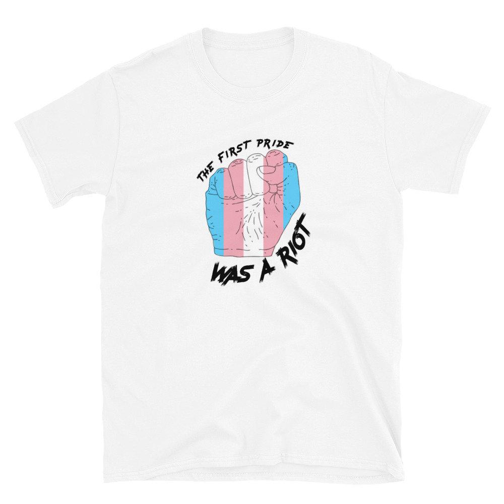 A white T-shirt features a illustrated black-and-white fist overlaid with the trans flag colors (light blue, pink, and white) in stripes and the words "The First Pride Was a Riot" in black scratched type, positioned above and below the fist.