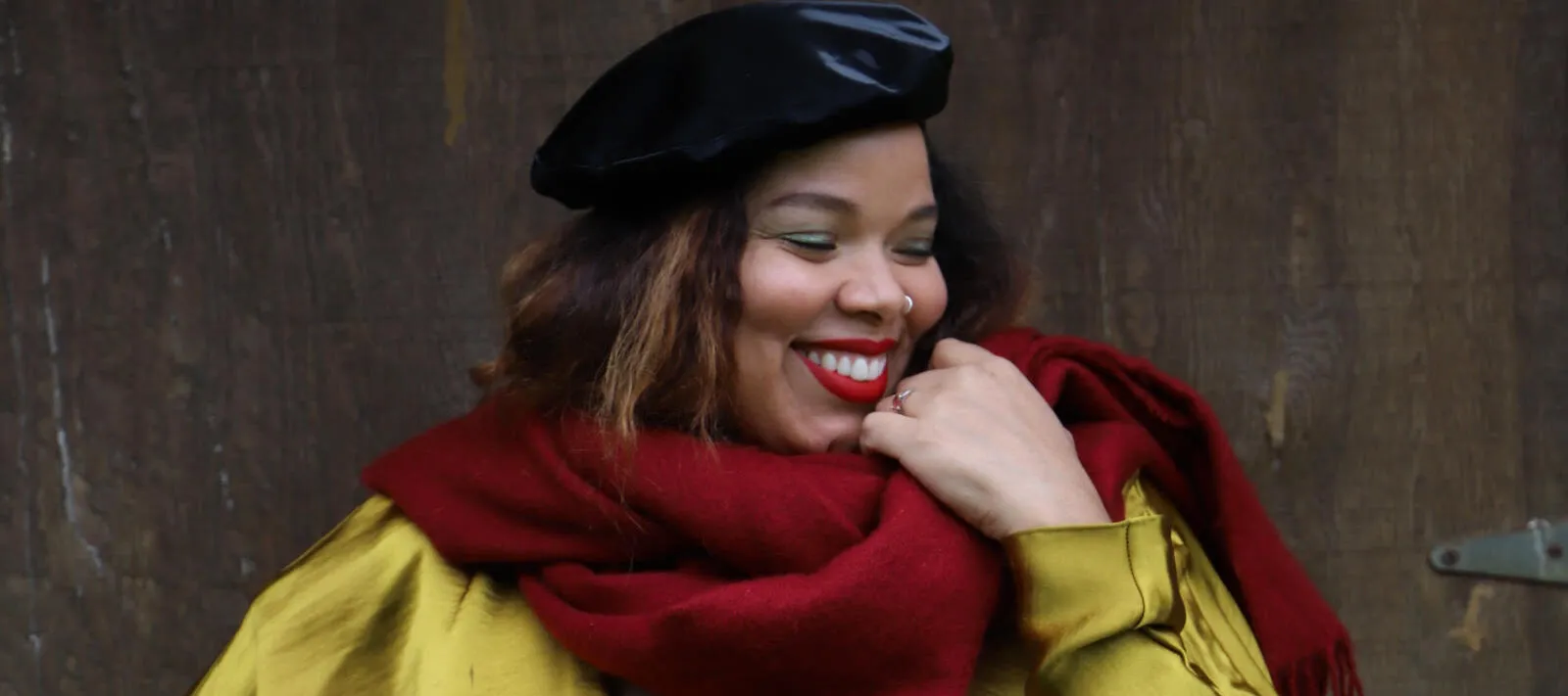 A medium-skinned woman stands in front of an industrial wall wearing a gold silk long sleeve dress with a deep V-neck, a black velour beret, and a cranberry red scarf wrapped loosely aroud her neck. With her right hand on her hip and her left brought up to the scarf, she smiles brightly, looking to the side.