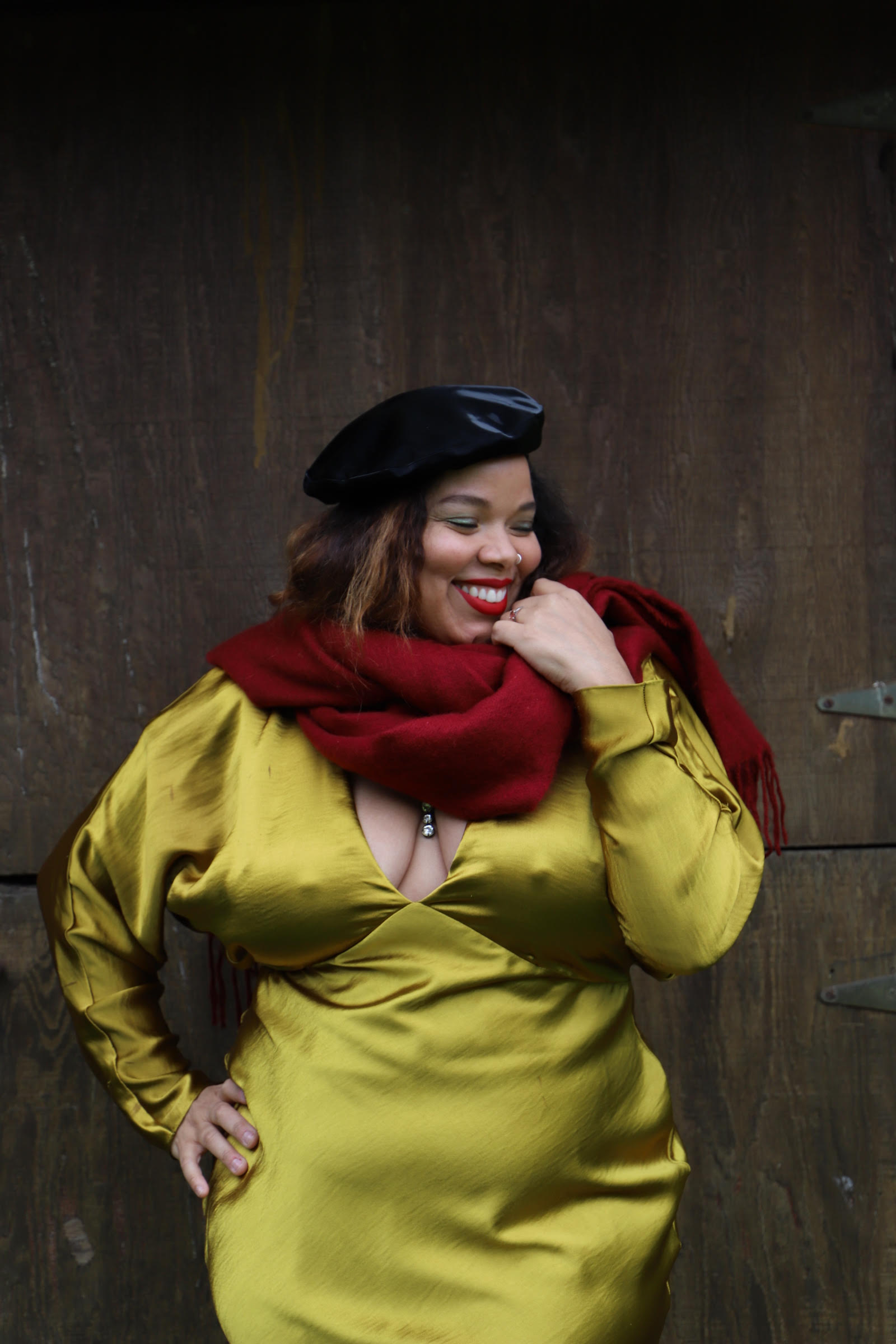A medium-skinned woman stands in front of an industrial wall wearing a gold silk long sleeve dress with a deep V-neck, a black velour beret, and a cranberry red scarf wrapped loosely aroud her neck. With her right hand on her hip and her left brought up to the scarf, she smiles brightly, looking to the side.