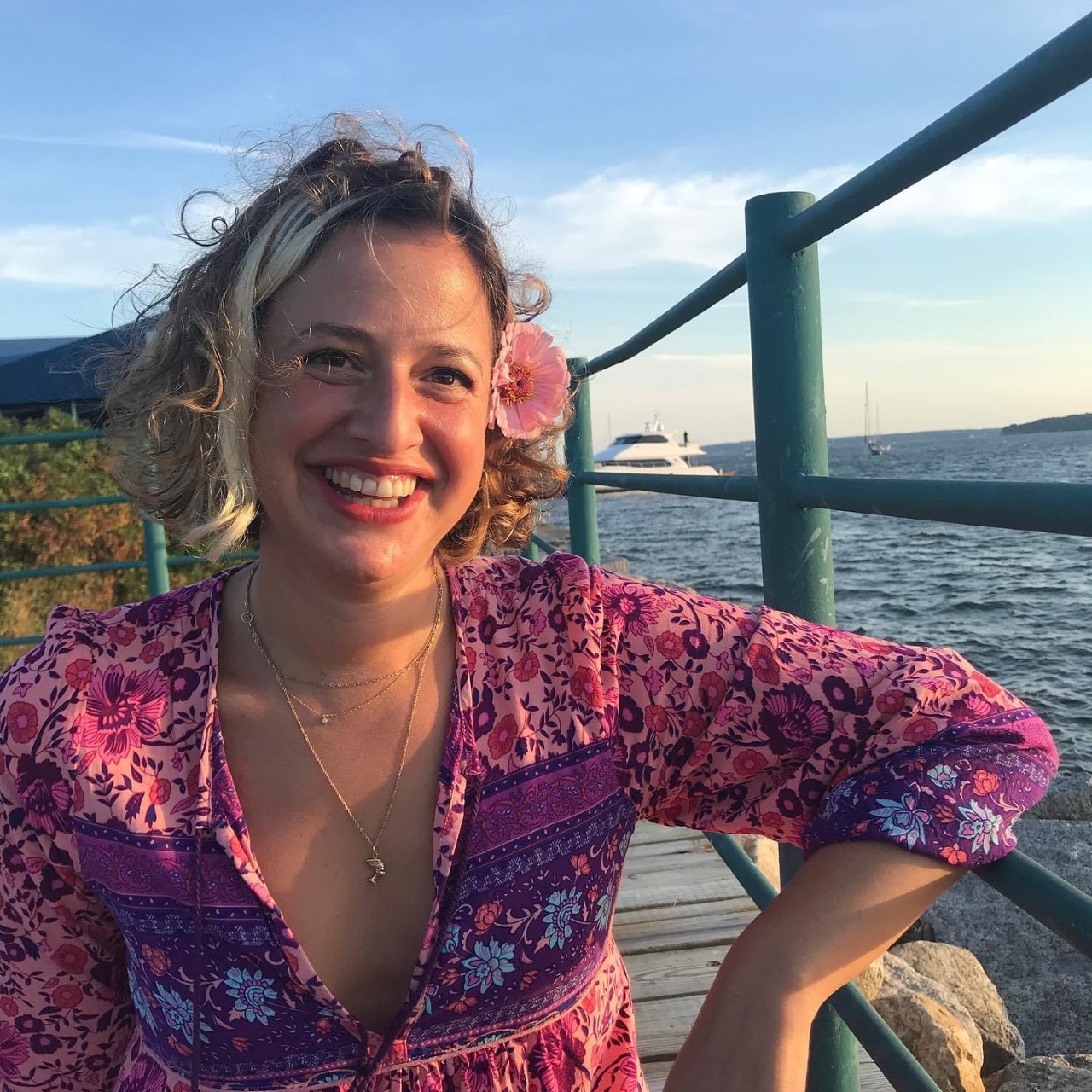 Sitting on a dock next to choppy water, a light-skinned woman smiles happily at the camera, her left arm resting on the dock railing. She wears a pink floral-patterned v-neck dress or shirt, and a pink blossom tucked behind her left ear. Her chin-length, light-brown hair is curly and blows in the wind.