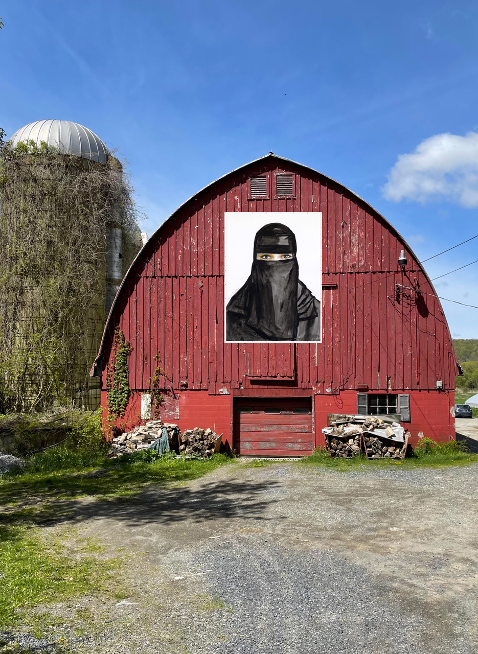 A red wooden barn stands at the end of a gravel lot, with a vine-covered silo behind it. On the barn is a large portrait photograph of a woman wearing a black burqa, only her eyes are revealed. The sky is bright blue and nearly cloudless.