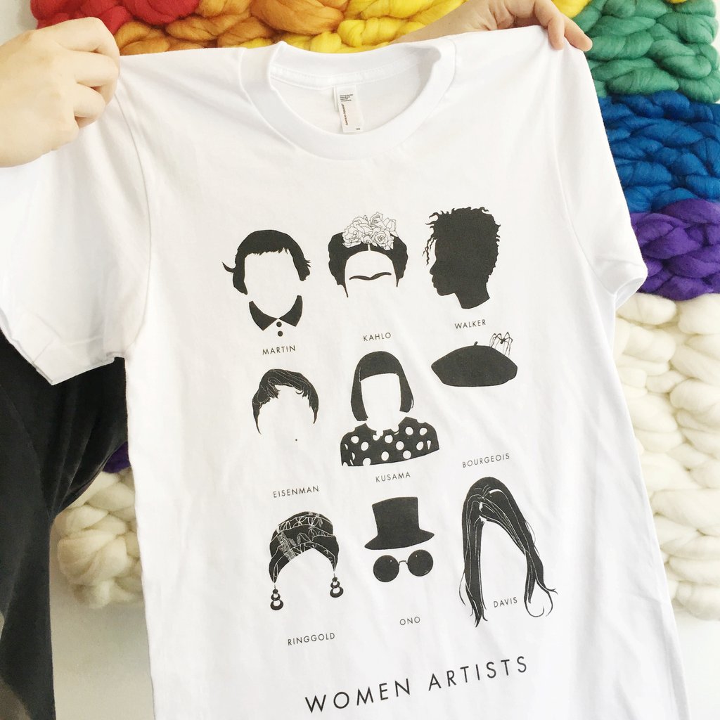 Two pale hands emerge from the left and hold up a white T-shirt featuring a grid of nine minamalist illustrations of the faces of famous women artists including Frida Kahlo, Kara Walker, Yayoi Kusama, Yoko Ono and more. At the bottom of the grid "Women Artists" is written in clean black type.