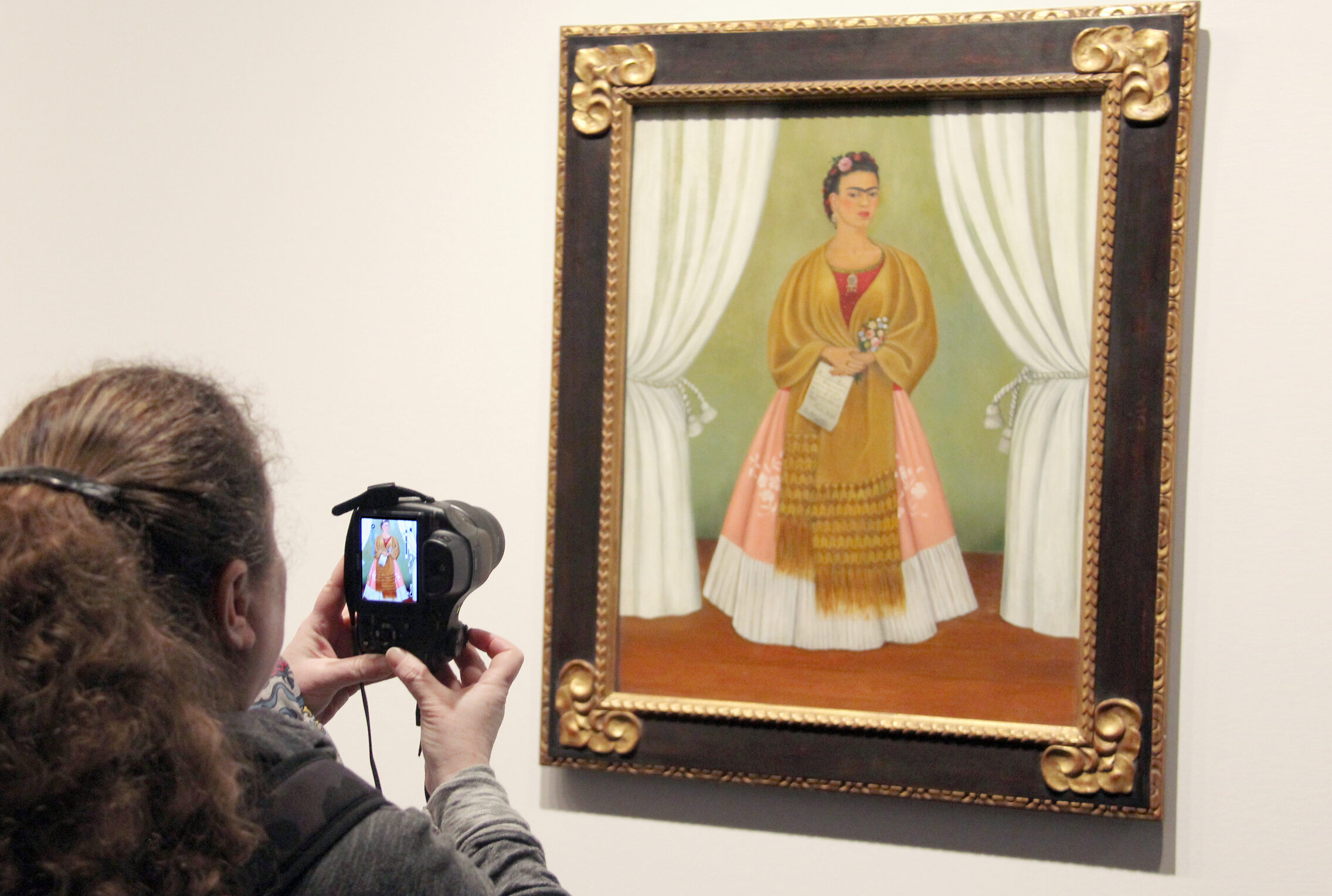 View from behind a light-skinned woman with long brown hair taking a photo of a painting of a medium-skinned woman in a dress in the museum galleries.