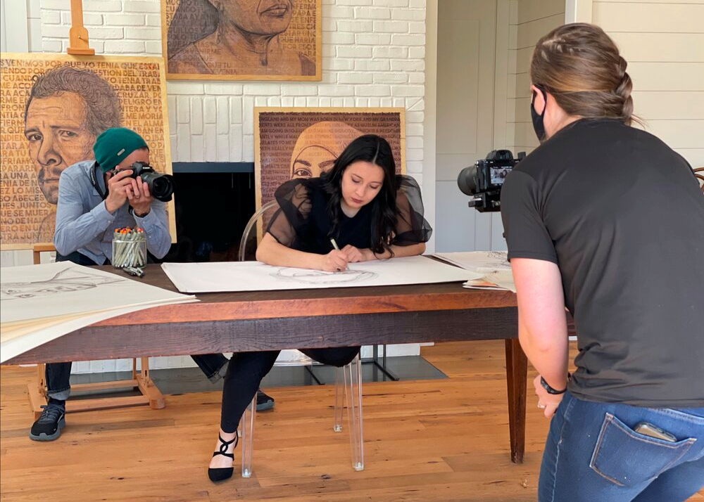 A light-skinned woman with long dark brown hair sits at a wooden table drawing a large face in pencil. A light skinned man with a camera stands behind her and a light skinned woman with a camera stands in front of her. Both figures are photographing her. Behind the artist are three of her finished portraits.