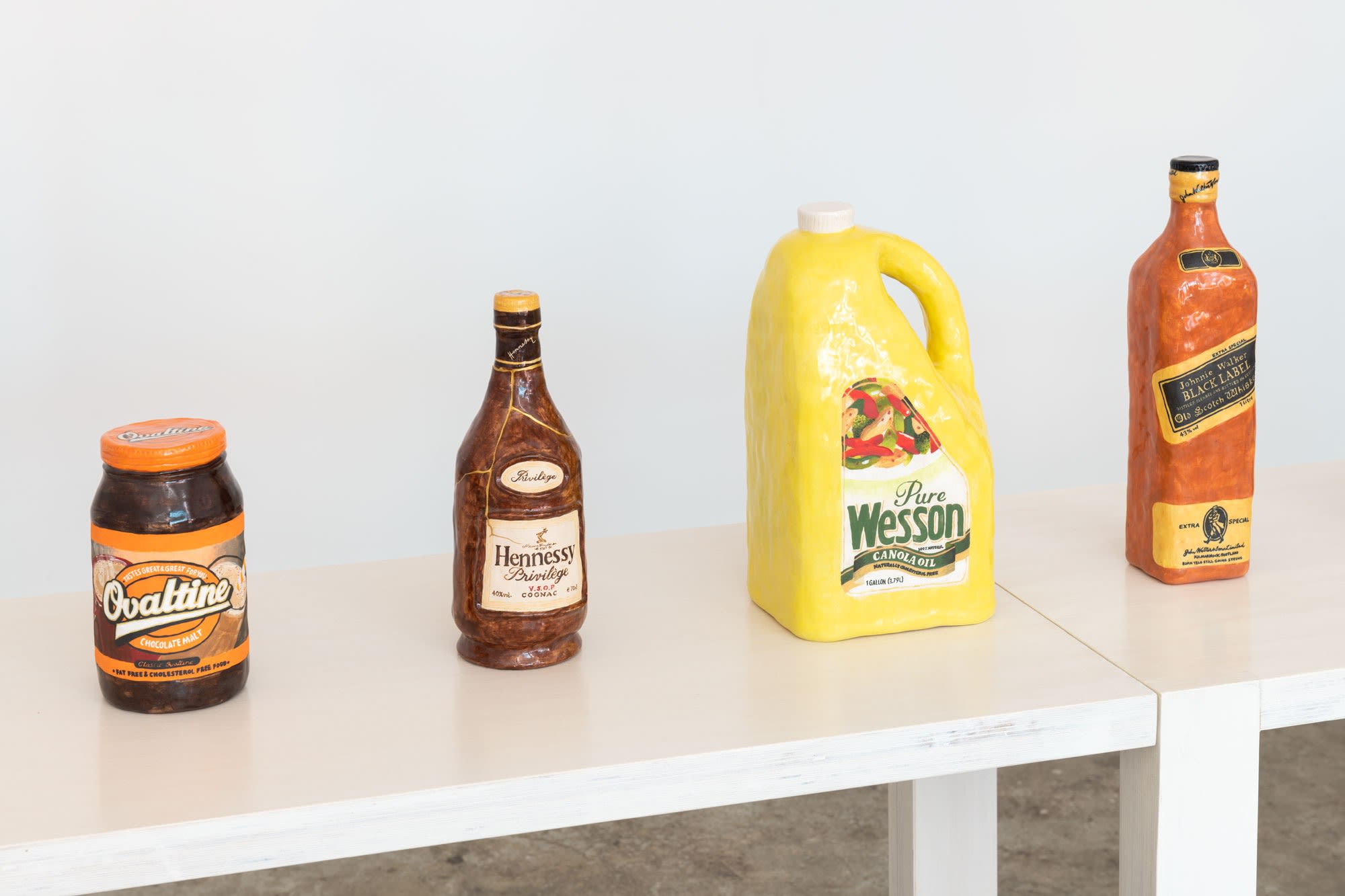 Six clay sculptures that look like grocery store products are arranged in a group. On the left, a can of condensed milk rests on top of a can of Spam. In the middle, a can of corned beef sits before a bottle of ketchup. On the right, a can of sweet corn is situated next to the ketchup bottle, while a package of Vienna sausages leans against the corned beef.