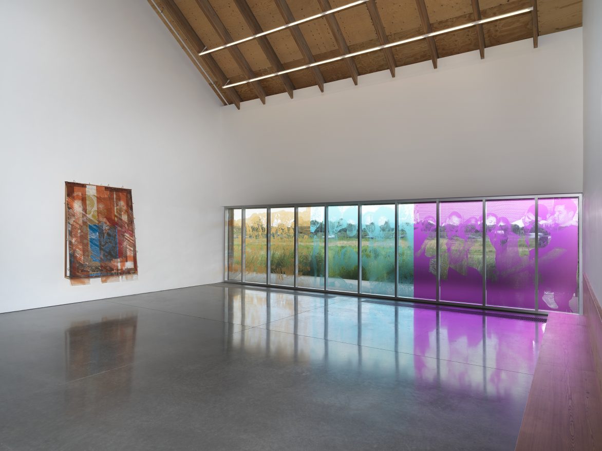An abstract mixed-media artwork hangs on the white wall of a large, modern gallery space. Next to it, a wall of windows is overlaid with yellow, light blue, and purple film that features images of people cut out in strategic spots. A field of waist-high grass is visible through the windows.
