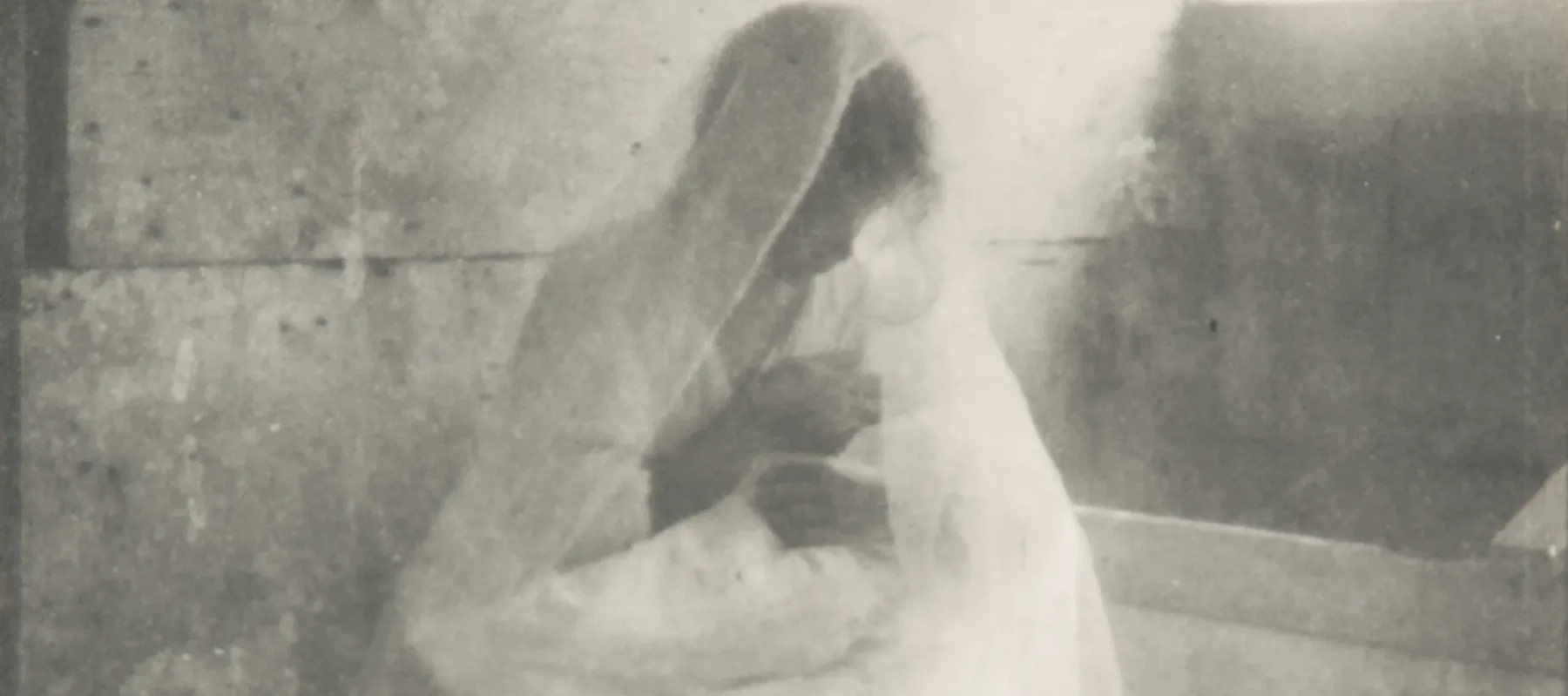 Ethereal black-and-white photograph of a women wearing a long white dress and gossamer veil, sitting in a stable, holding a swaddled infant. Dramatically illuminated by a shaft of light streaming in, she gazes down at the child cradled in her arms.