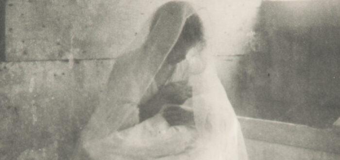 Ethereal black-and-white photograph of a women wearing a long white dress and gossamer veil, sitting in a stable, holding a swaddled infant. Dramatically illuminated by a shaft of light streaming in, she gazes down at the child cradled in her arms.