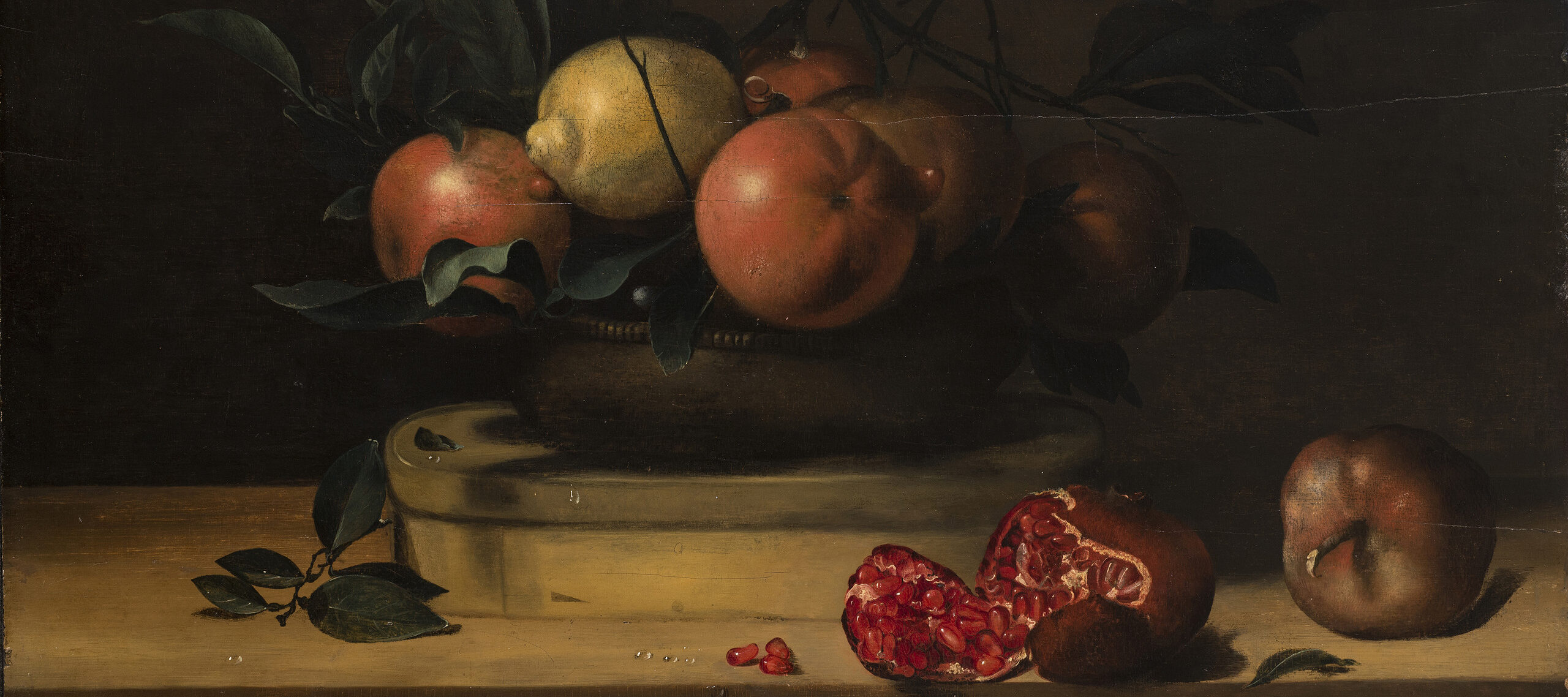 A vessel mounded with 5 oranges, a lemon, and greenery sits atop an oval, wooden box resting on a wooden plank. In the foreground, two pomegranates, one of which has split open and dropped three seeds, balance near the plank’s edge. Droplets of water dot the fruit and the table.
