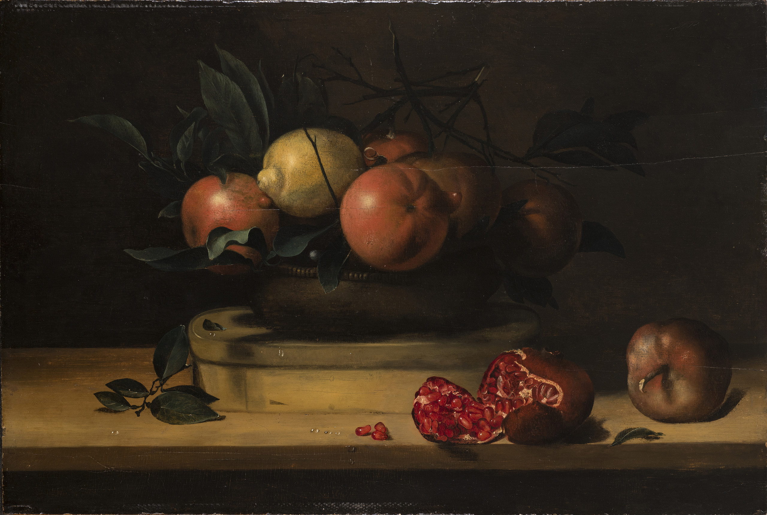 A vessel mounded with 5 oranges, a lemon, and greenery sits atop an oval, wooden box resting on a wooden plank. In the foreground, two pomegranates, one of which has split open and dropped three seeds, balance near the plank’s edge. Droplets of water dot the fruit and the table.